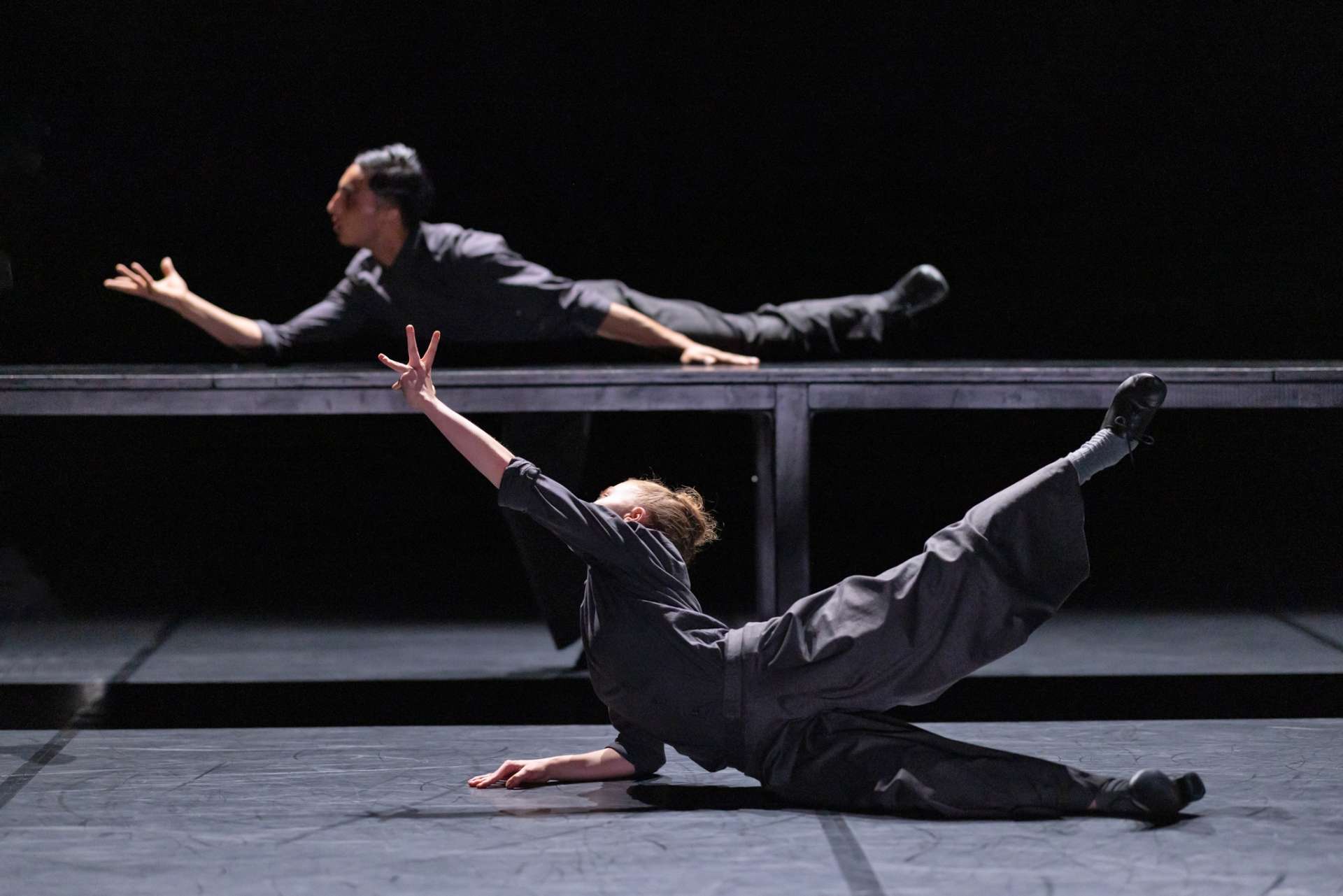 Deux interprètes de la pièce Cathedral sont allongés au sol et sur une table. L'atmosphère est noire tout comme leurs tenues. 