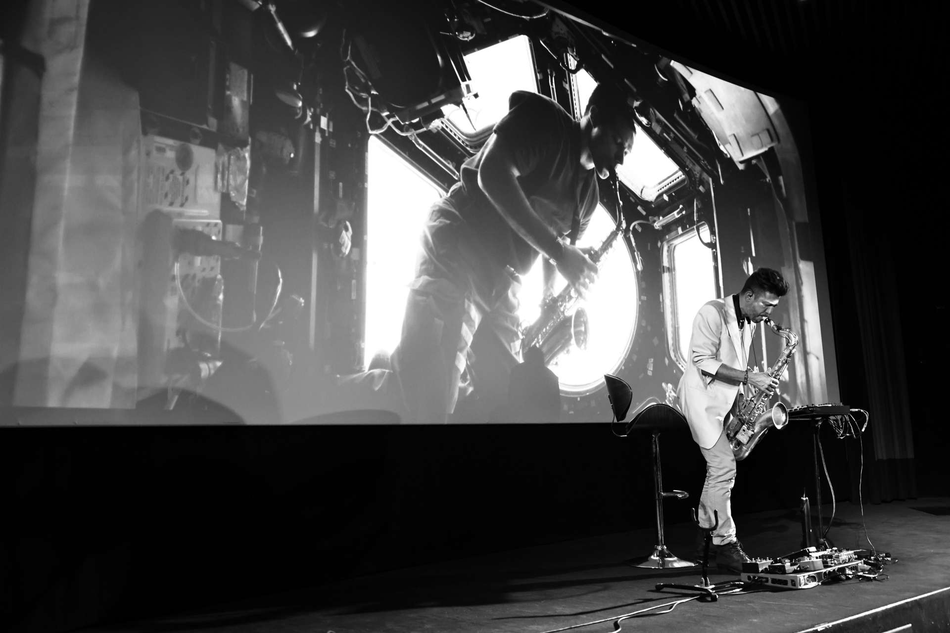 Photo en noir et blanc du saxophoniste Guillaume Perret jouant de son instrument sur scène. Derrière lui, une écran géant projette une image de Thomas Pesquet jouant également de saxophone dans la station spatiale internationale.