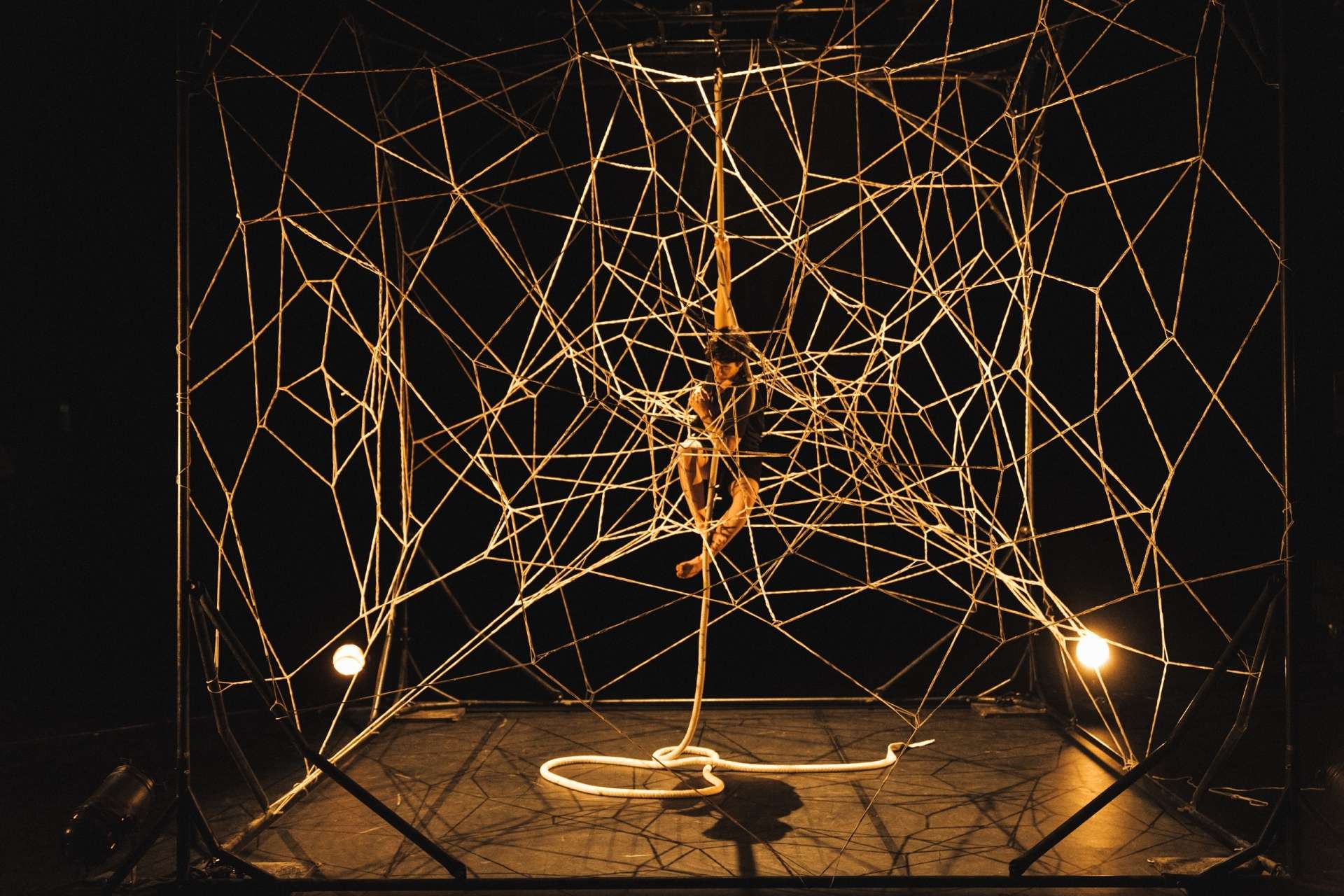 Photo de Inbal Ben Haim, circassienne, suspendue à l’intérieur d’une grande structure qui ressemble à une toile d’araignée géante. La toile est faite de nombreux fils fins, étirés entre un cadre métallique noir. Deux projeteurs sur le sol de la scène diffuse une lumière vive, contrastant avec l'arrière plan noir. 