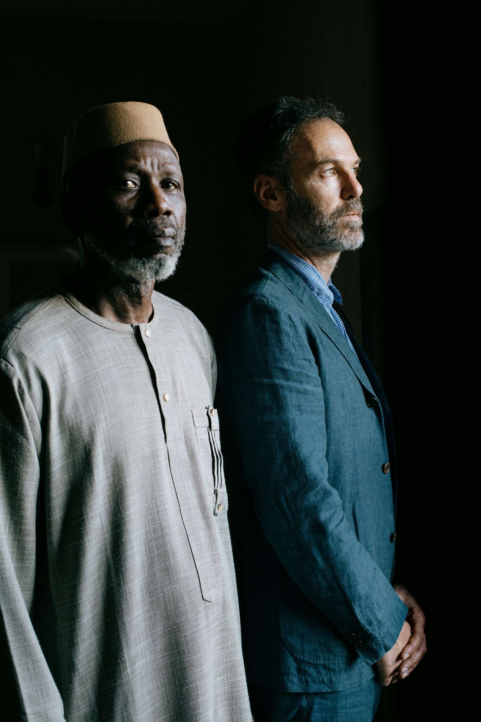 Portrait de Ballaké Sissoko, au premier plan, et Piers Faccini, au deuxième plan. Les deux sont de profil. Ballaké Sissoko porte une longue chemise grise et un chapeau beige. Piers Faccini porte une chemise et une veste bleues. 