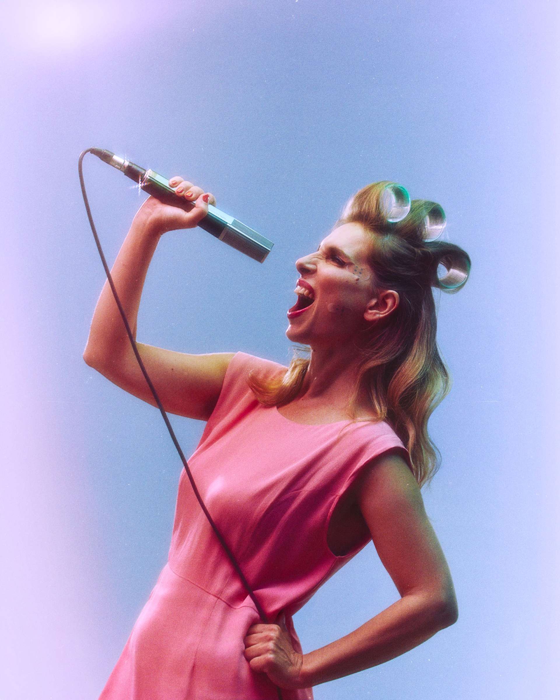 Photo portrait de profil de Barbara Carlotti style rétro, habillée d'une robe rose, se penchant en arrière elle tient dans sa main un micro filaire et chante. Ses cheveux blond, sont tenues par des bigoudis.