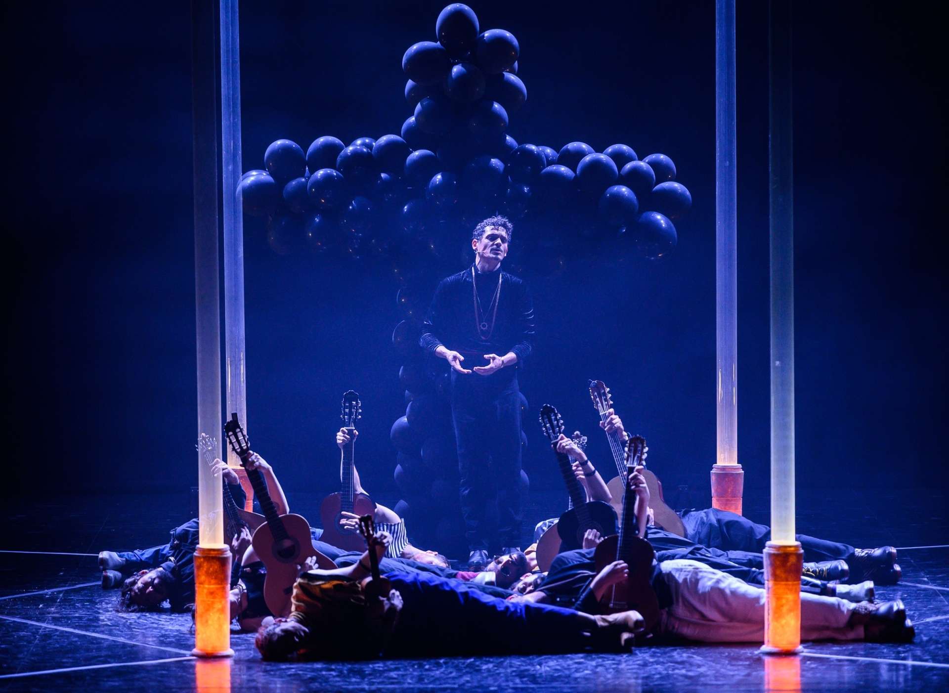 Un chanteur de fado debout au centre d’un cercle de danseurs allongés, guitare au sol, en arrière plan, une grande croix formée de ballons.