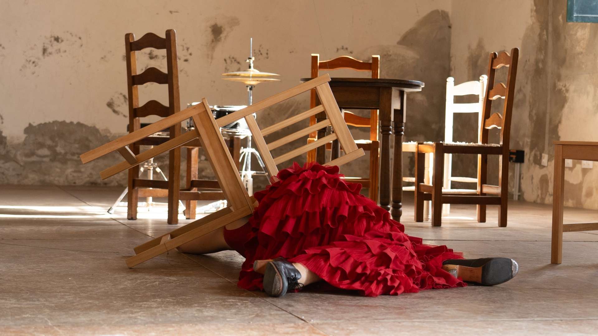 Dans une pièce aux murs délabrés, une danseuse de flamenco allongée par terre. Elle est prise au milieu de chaises en bois. On ne voit que sa robe à volants traditionnelle rouge et ses chaussures noire. Un charleston et une caisse claire au deuxième plan parmi les chaises.