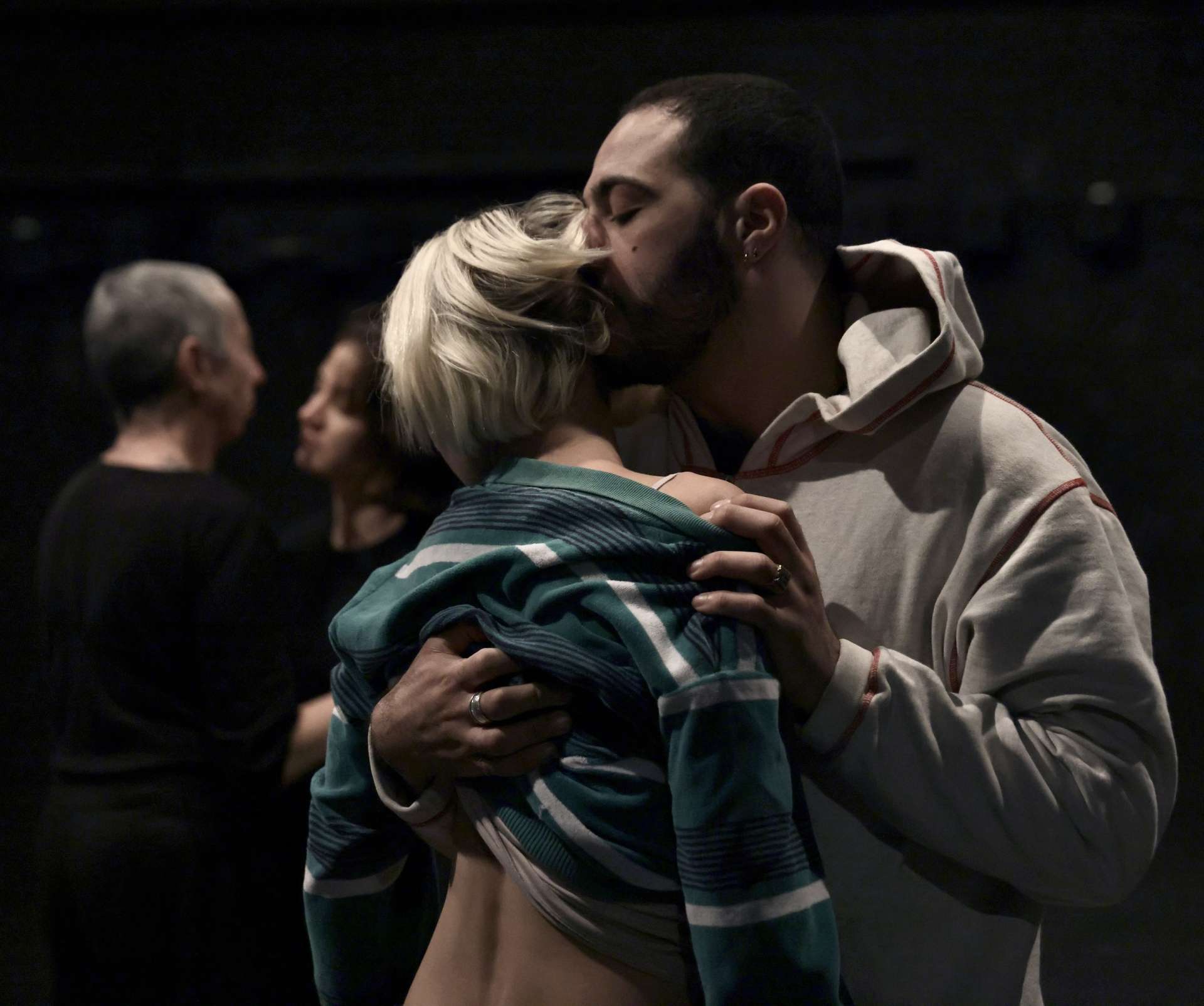 L'image montre un homme enlaçant une femme. La femme nous tourne le dos, on peut apercevoir une parcelle de sa peau, l'homme soulevant légèrement son haut. Des personnes sont également présentes, floues en arrière-plan. L'homme porte un sweat à capuche clair.