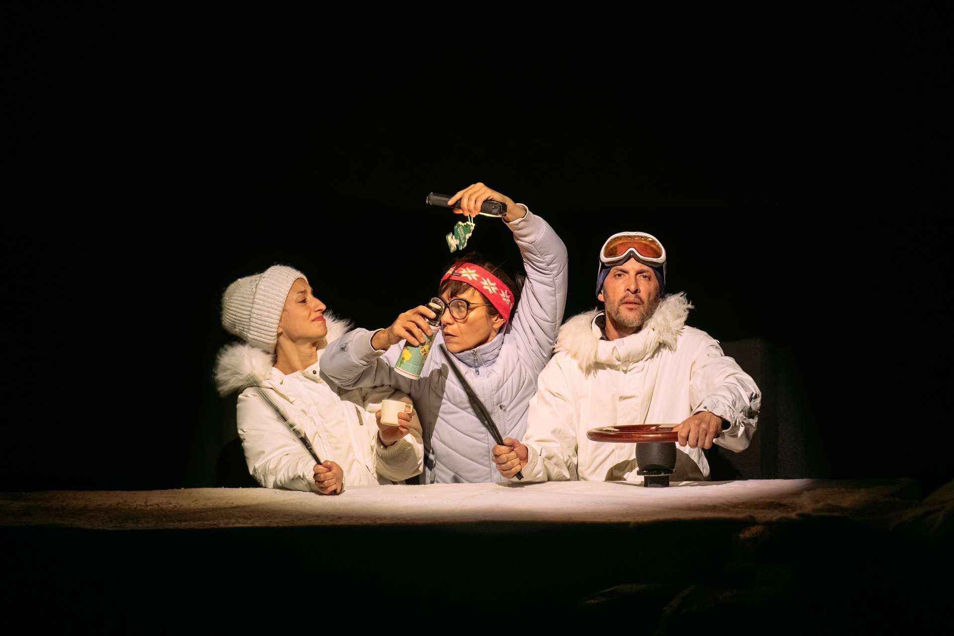 Une scène théâtrale met en scène trois personnages vêtus de vêtements blancs d'hiver. Deux femmes, l'une portant une casquette et une autre un bandeau avec un motif de Noël, ainsi qu'un homme, avec un masque de ski. Les personnages sont engagés dans des actions, comme tenir des objets, gesticuler, et se positionner de façon à suggérer une interaction. 