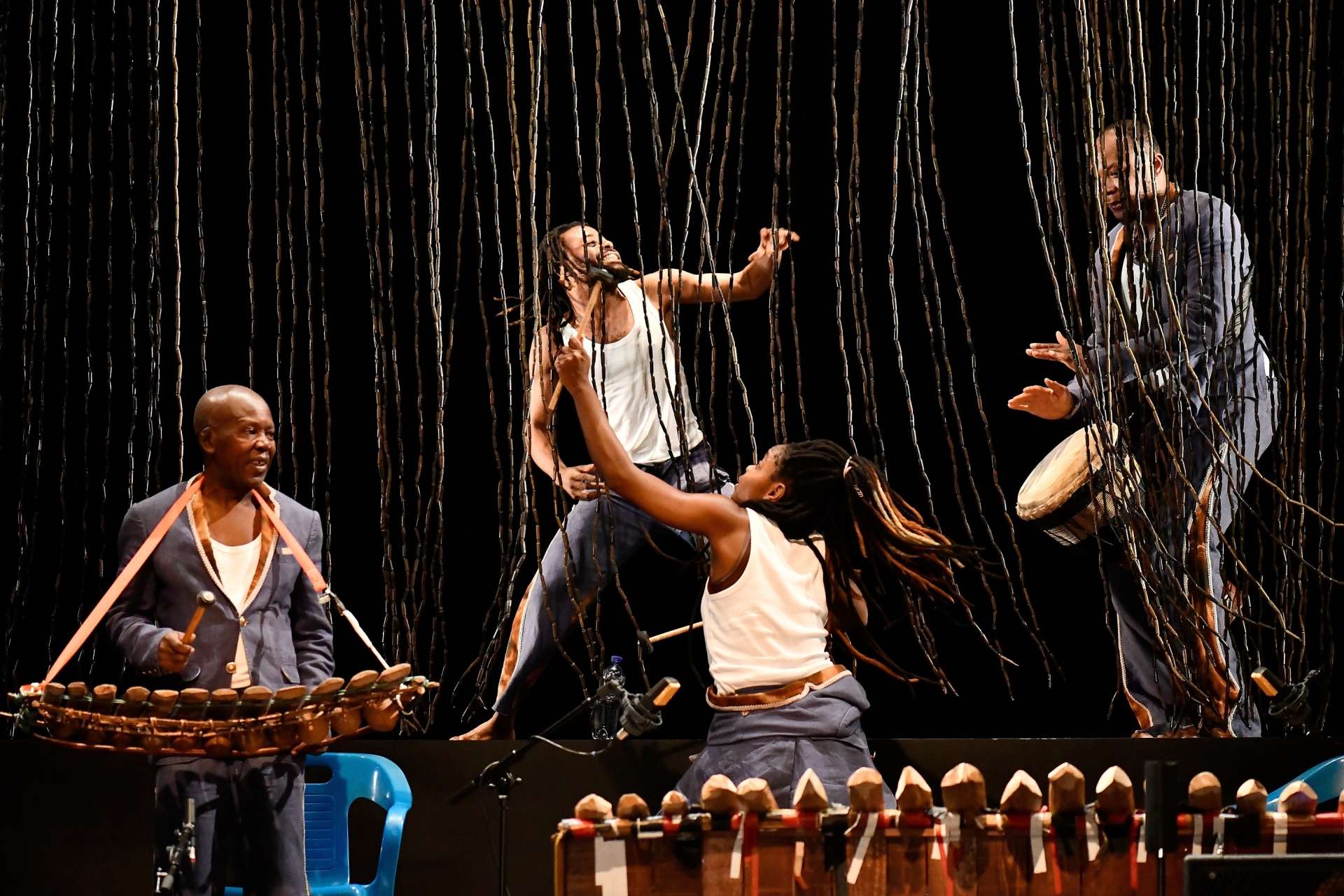 Quatre musiciens sont en plein jeu, chacun à son instrument (balafon, tambour africain...). L'arrière-plan est une toile de fond sombre. Les musiciens sont séparés du premier plan au deuxième plan par un rideau de tiges verticales minces et souples qui tombent du plafond.