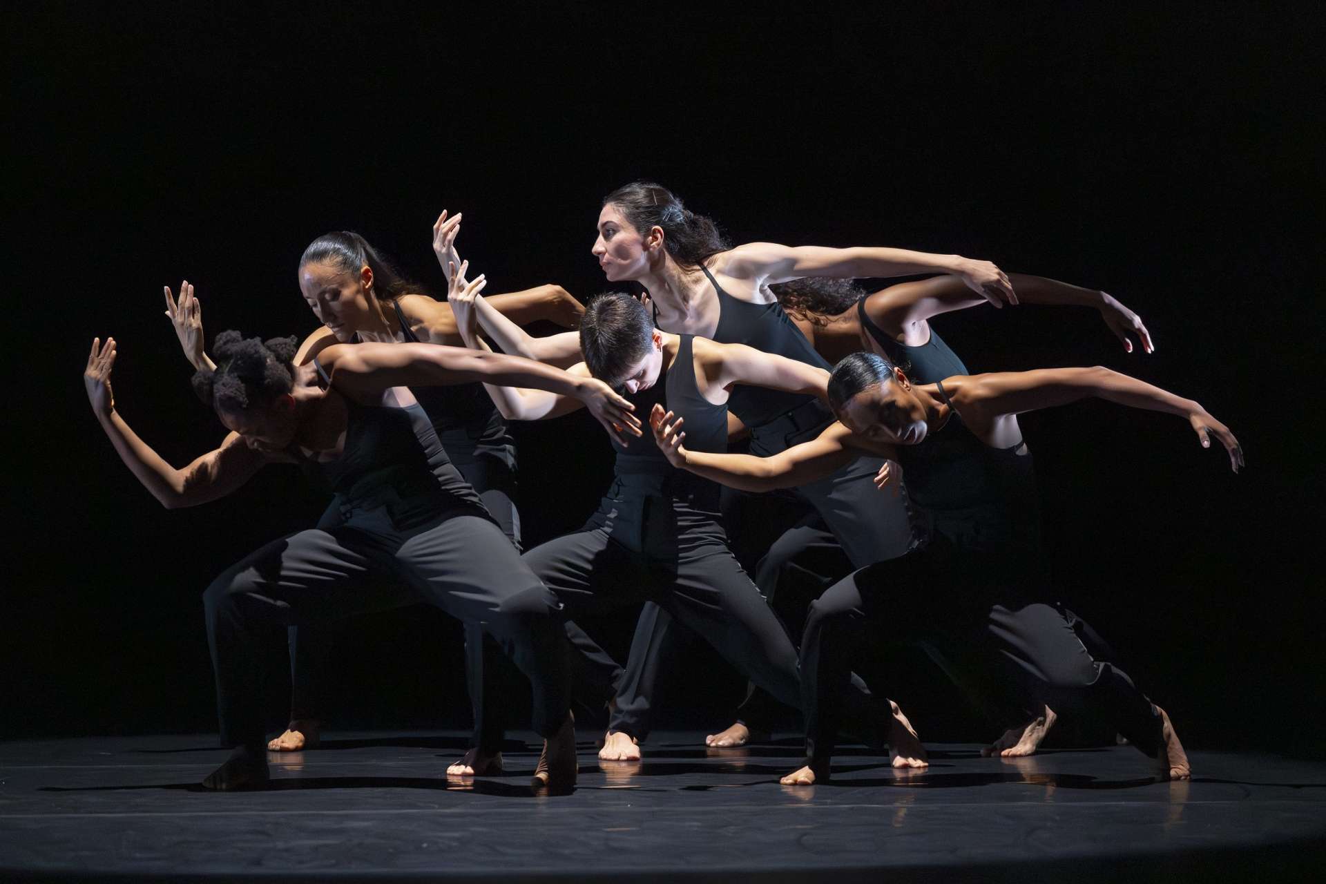 Photo d'un collectif de danseuses vêtues de noir qui exécute une chorégraphie de danse moderne jouant en synchronisation sur un fond noir. La chorégraphie met l'accent sur des mouvements de bras fluides et étendus. 