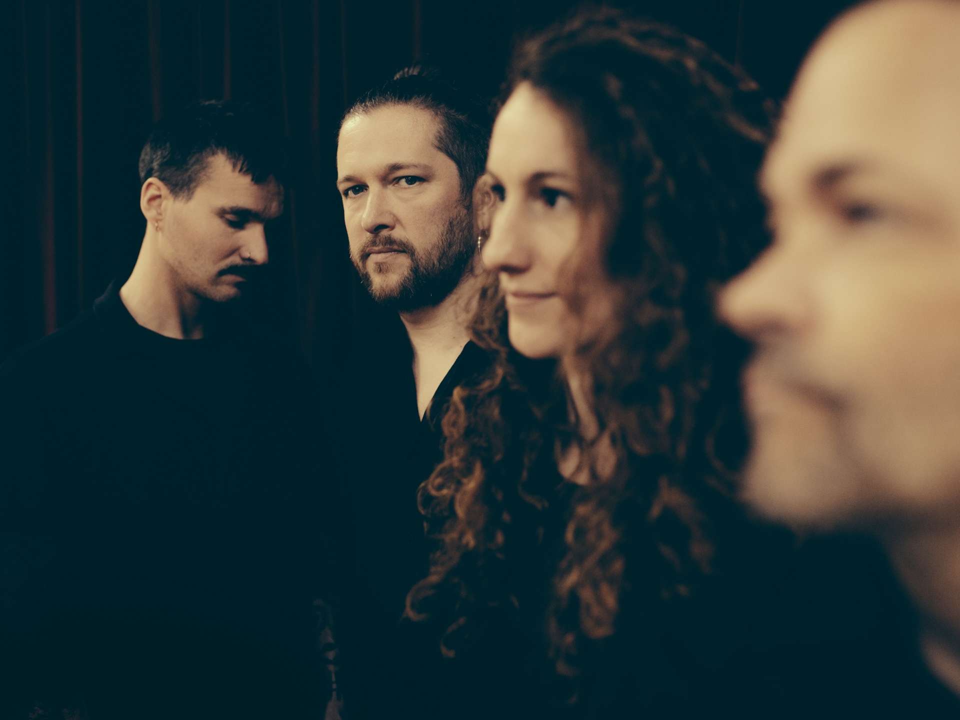 Portrait des quatre membres du groupe. Trois hommes, une femme. Ensemble sombre, sauf les visages qui ressortent. La photo joue sur la profondeur de champ.