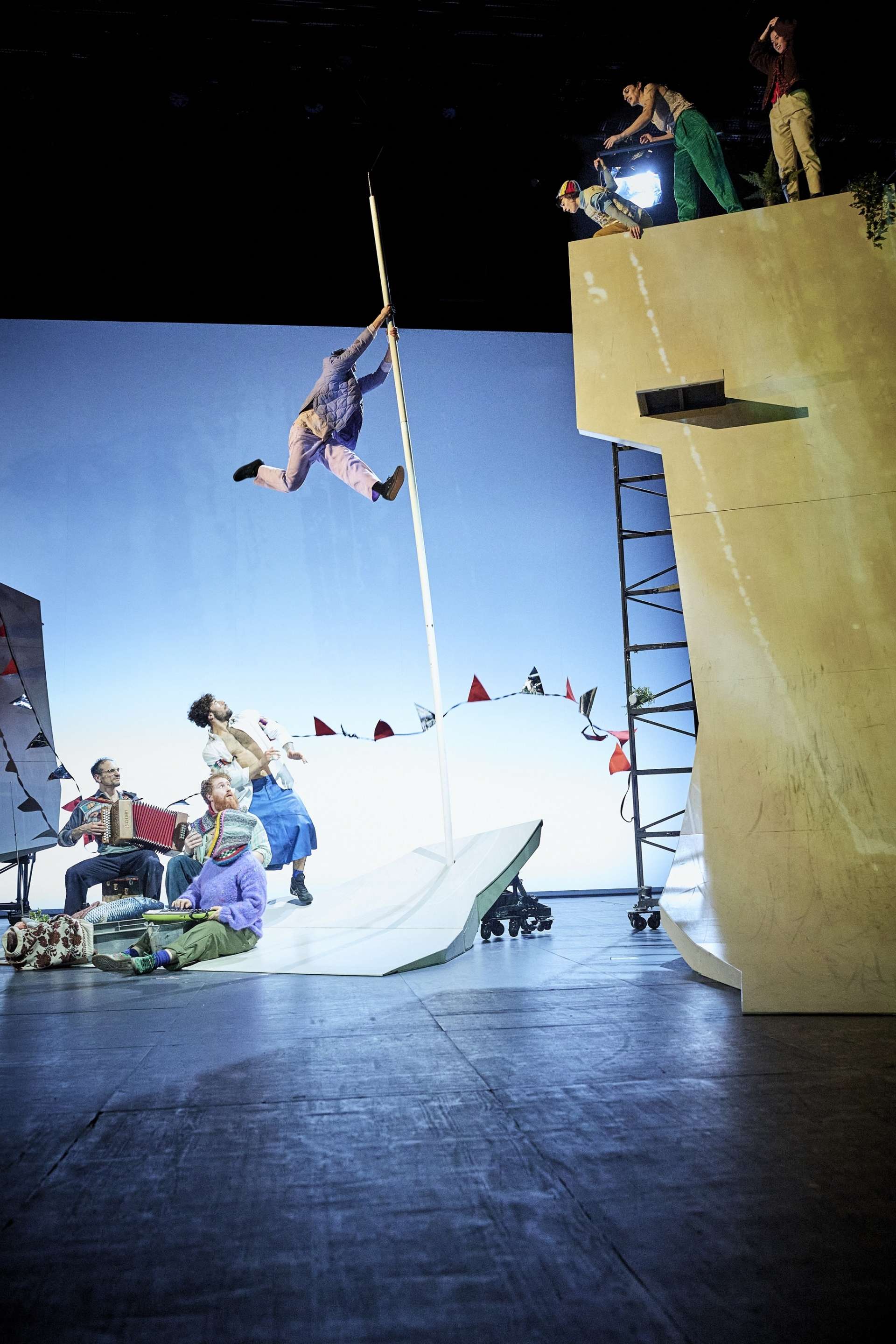 Un interprète s’élance en l’air accroché à une perche verticale. En haut d’une structure, quatre personnes observent la scène, tandis qu’au sol, un petit groupe joue de la musique. Le décor est urbain et festif : guirlandes, instruments...)