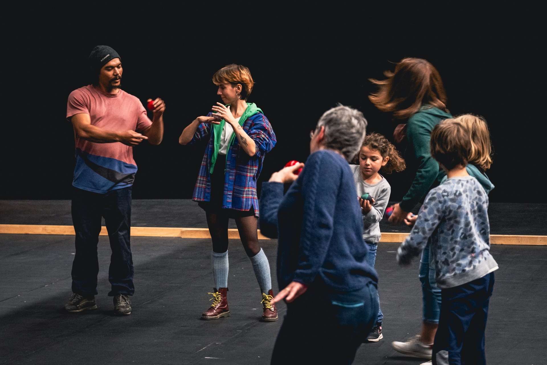 Image d'un atelier de jonglage avec un artiste du spectacle "Radio Maniok" (saison 24/25), autour de lui, des personnes de tout âge, tente de reproduire ses gestes, et de garder une boule de jonglage en équilibre sur le dos de leurs mains.