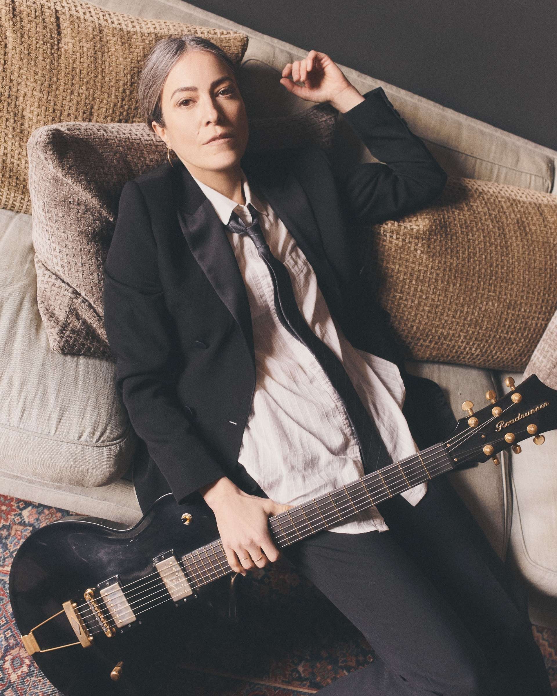 Photo de Keren Ann prise du dessus. Elle est allongée confortablement sur des coussins clairs. Elle tient une guitare électrique noire dans sa main droite et regarde en direction de l'objectif. Elle porte une chemise beige, une cravate, une veste et un pantalon noirs.