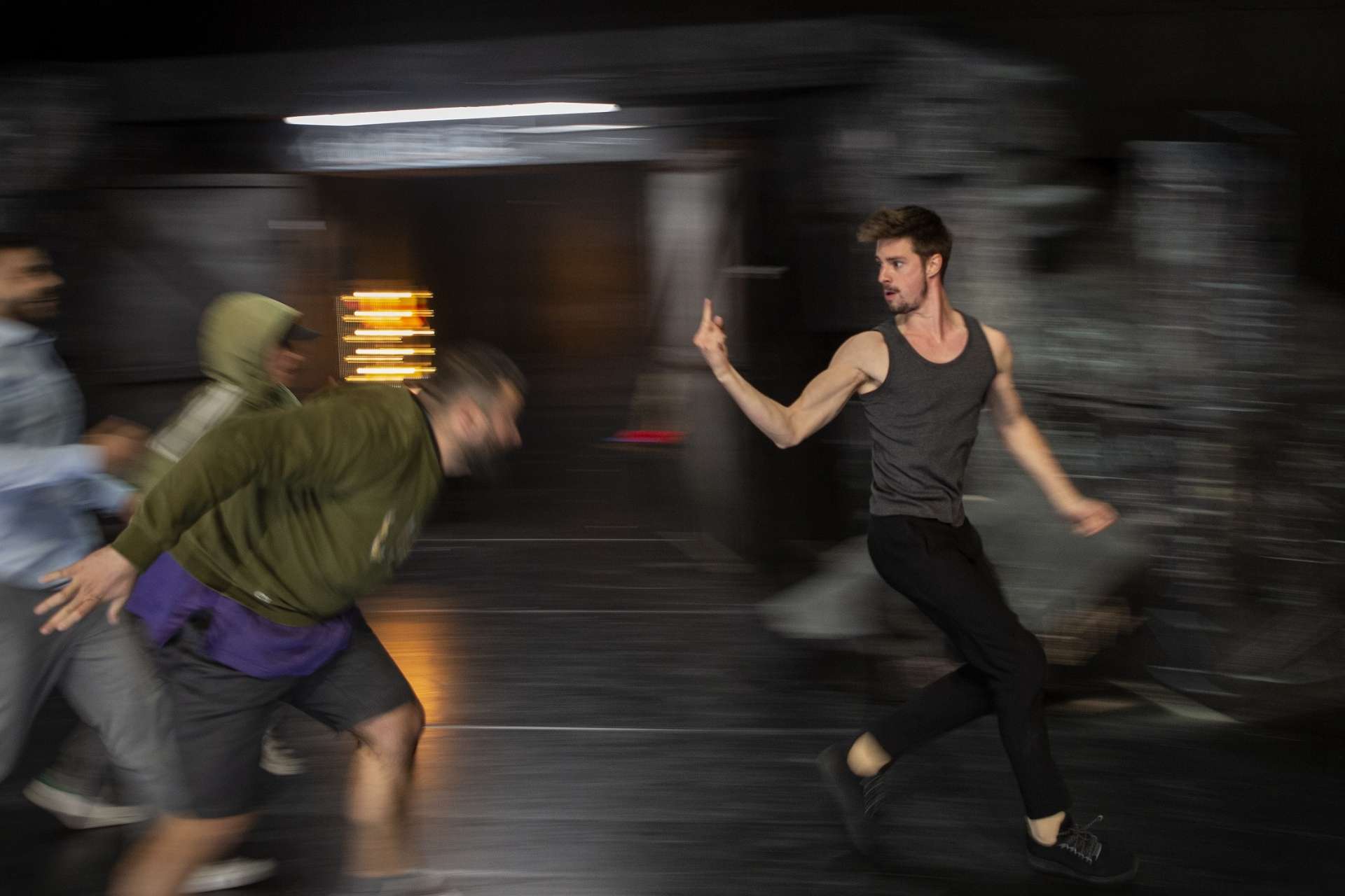 Dans un effet de mouvement rendu par du flou, un jeune homme en marcel kaki fait un doigt d'honneur à trois autres jeunes hommes qui le pousuivent.