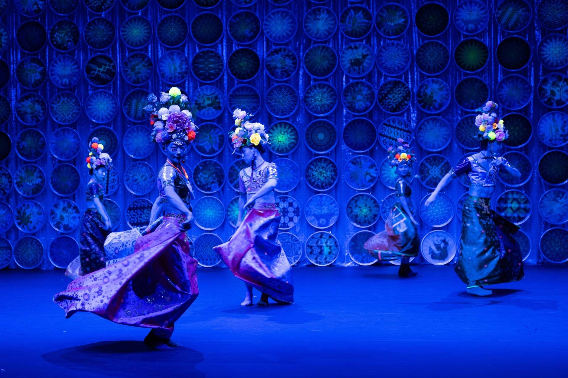 Plusieurs danseuses, vêtues de robes colorées et de coiffes décorées de fleurs, dansent sur une scène illuminée de lumière bleue.