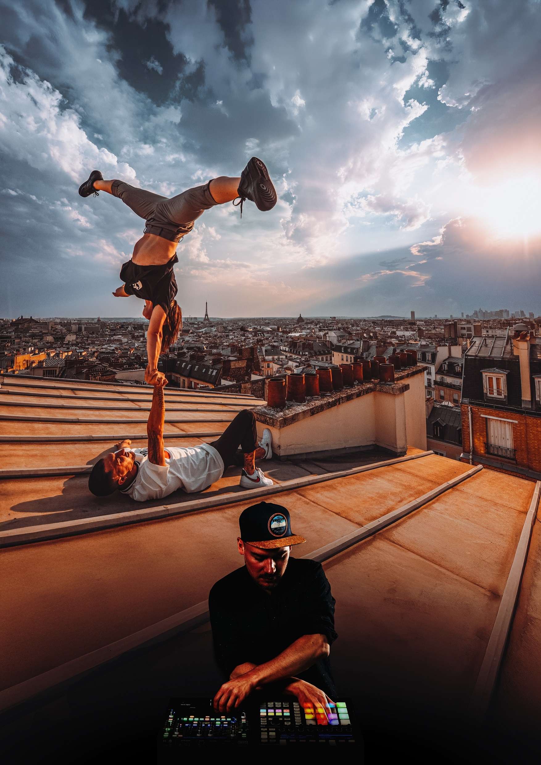 Photo montage, sur les toits de Paris deux acrobates : l'un allongé sur le sol un bras en l'air main dans la main avec l'autre acrobate en équilibre, réalisant un poirier sur un bras, les jambes écartées. Au premier plan, un dj sur une table de mixage.