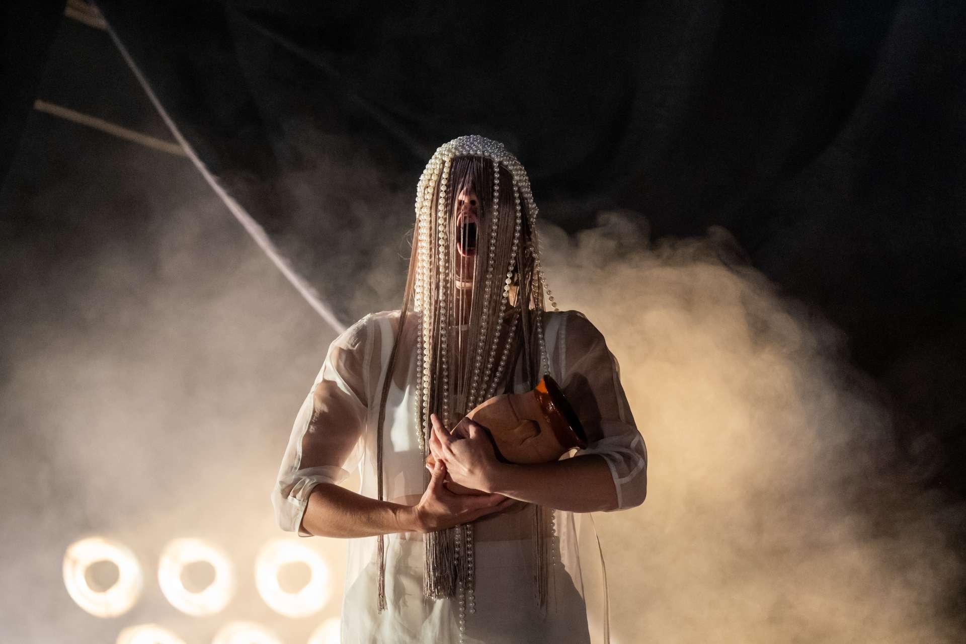 Une femme aux longs cheveux ornés de perles, portant une robe blanche et tenant un vase brun. Elle se tient devant un arrière-plan de fumée et de lumières. La bouche ouverte, elle semble chanter ou avoir peur.