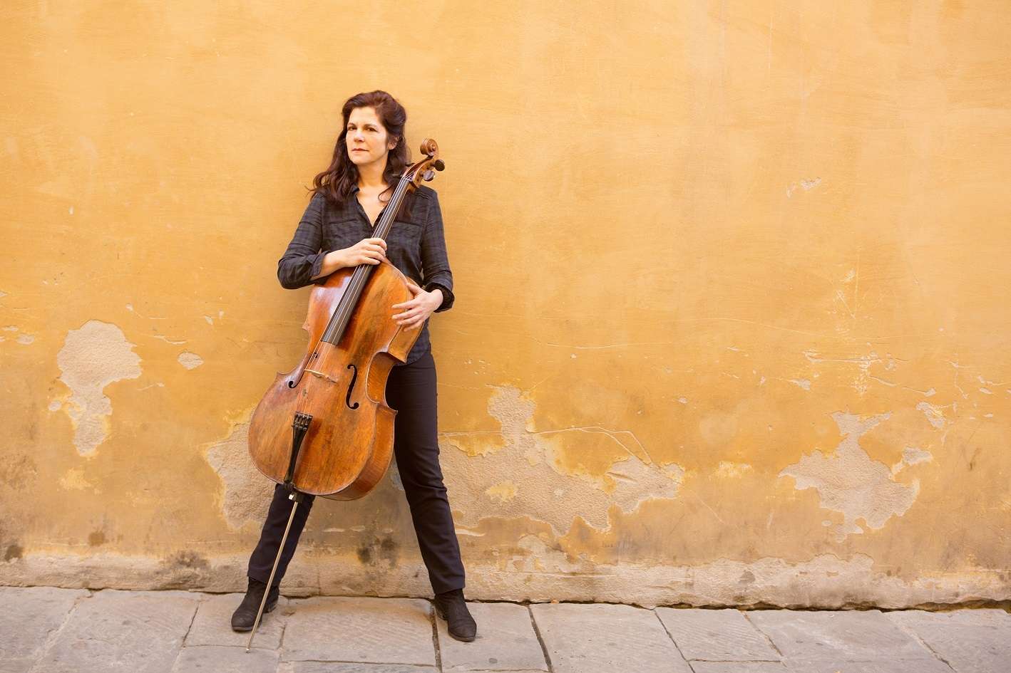 Photo de Sonia Wieder Atherton devant un mur jaune, un petit peu abîmé. Elle se tient debout avec, devant elle, son violoncelle.