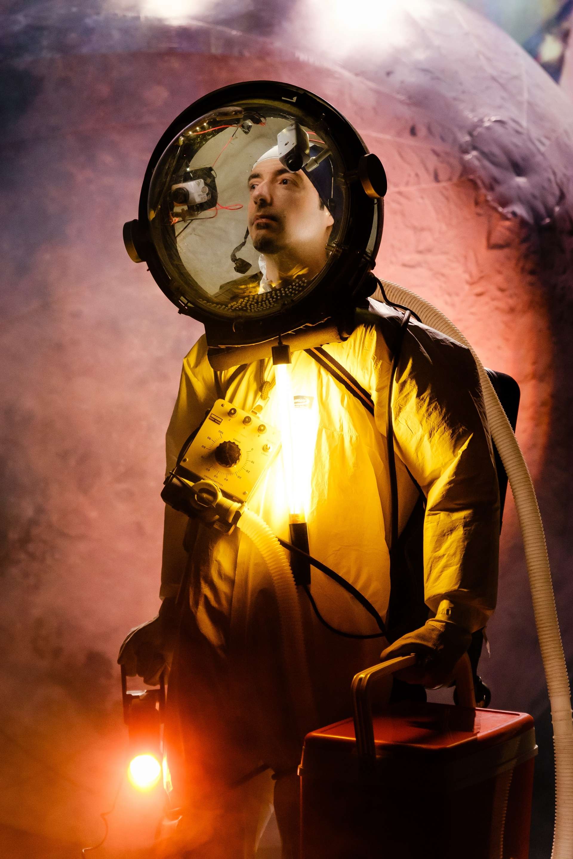 L’image présente un astronaute en gros plan, vêtu d’une combinaison spatiale jaune vif et de plusieurs accessoires. Le casque de l’astronaute est transparent et sphérique, permettant de voir son visage. On peut observer de petites caméras fixées à l’intérieur du casque. L’arrière-plan est flou mais montre une sphère ressemblant à la surface d’une planète.