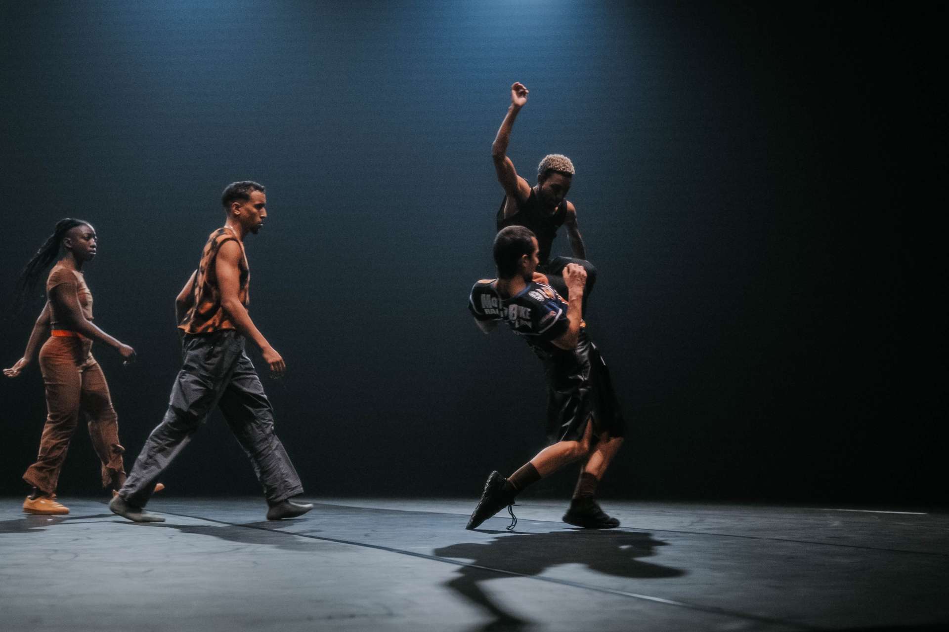 L'image montre quatre personnes, chacune engagée dans des mouvements distincts. Au centre, à droite de l'image, deux artistes interagissent. L'un semble être en hauteur, soulevé en l'air par l'autre et un bras levé, ajoutant du dynamisme. A gauche de l'image, deux personnes sont en pleine marche. 