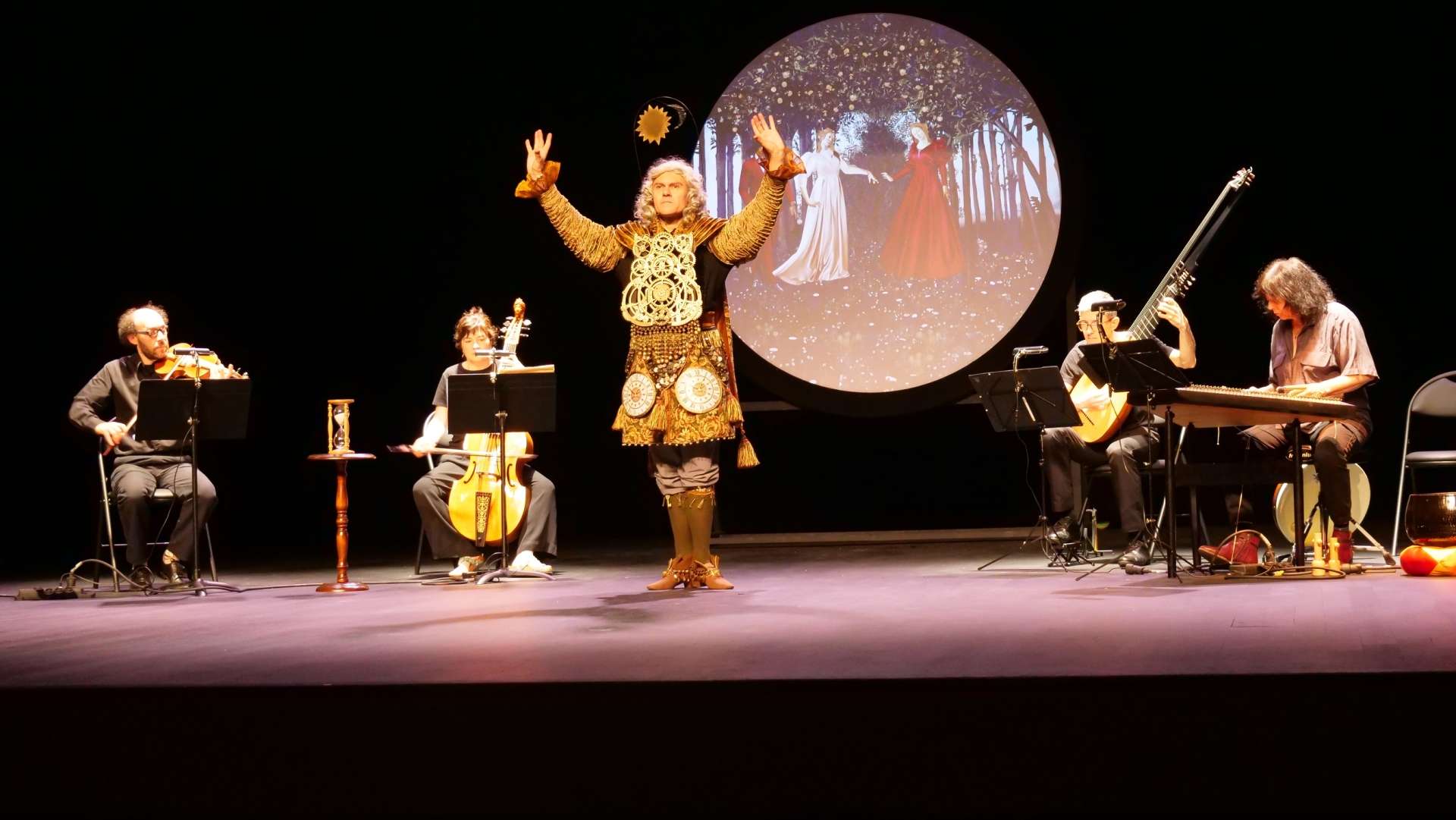 La scène est composée d'un orchestre disposé autour du personnage principal du spectacle "Tempus Fugit". Celui-ci est en pleine performance, ses bras levés, vêtu d'une tenue ornée de détails dorés, ressemblant à un costume de Louis XIV, le Roi-Soleil. Les musiciens et musiciennes qui l'entourent sont en position assise, jouant d'instruments (violon, viole de gambe...).