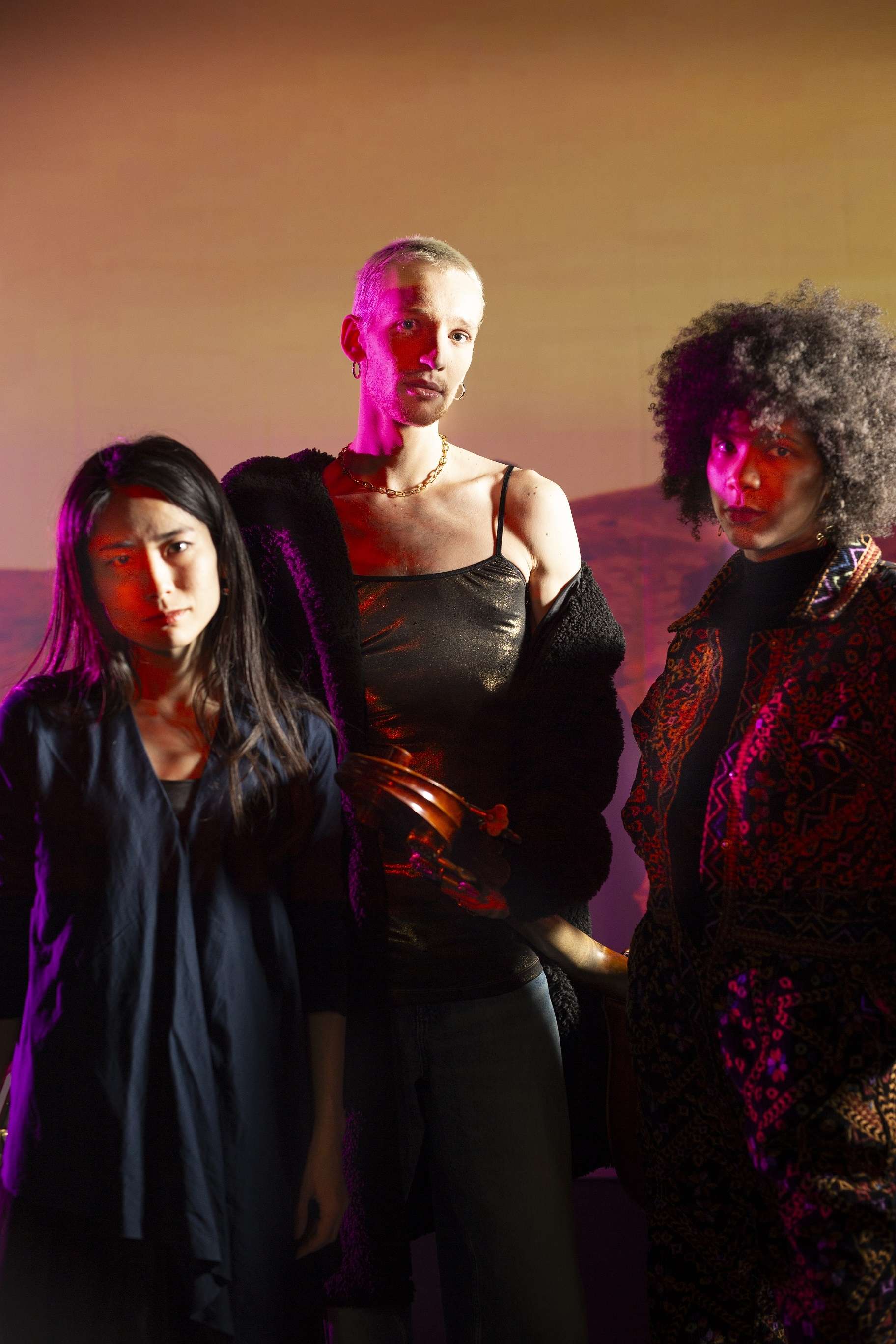 Photo portrait des trois artistes de "The Democracy Project". Derrière un arrière plan qui semble un écran projetant un paysage de montage ou de dune de sable. Une lumière rose fluo reflète sur le visage des interprètes. 