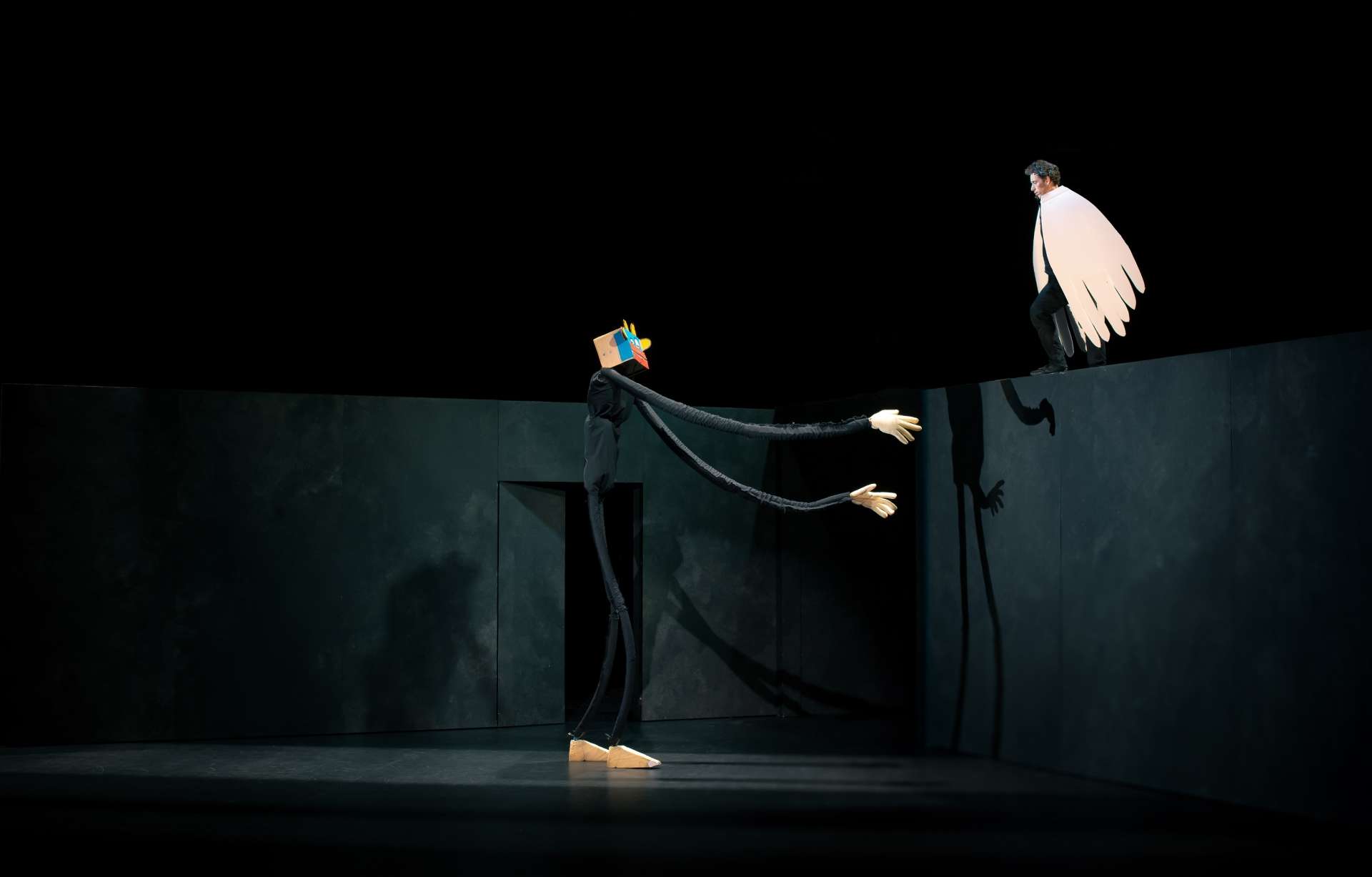 Une scène de magie nouvelle, met en scène deux personnages : Le premier est une figure allongée avec de longues membres et porte un masque sur la tête composé d'une forme géométrique colorée, tandis que le second personnage est un être humain avec une cape blanche en forme d'ailes, semblant s'élever au-dessus d'un mur.