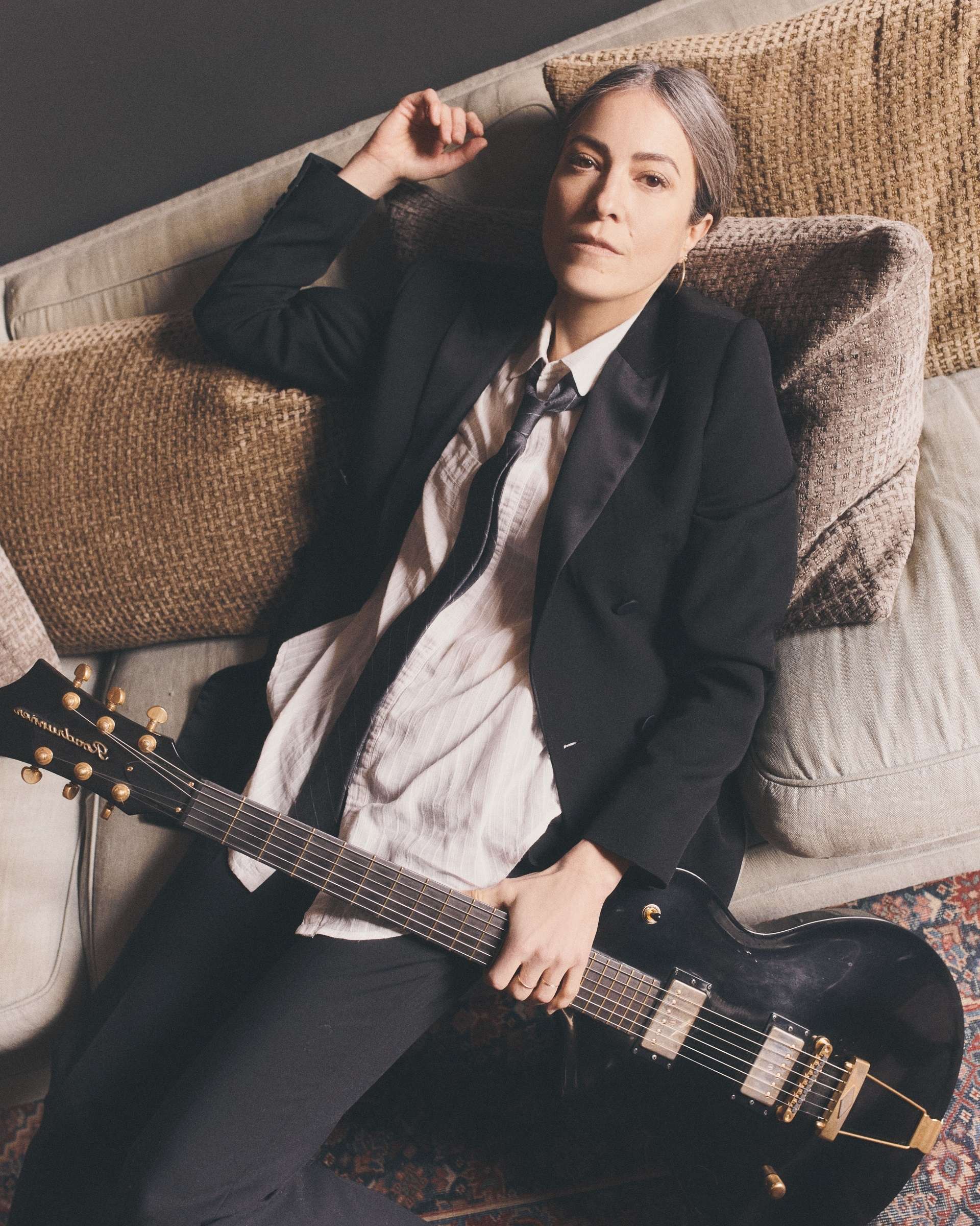 Photo de Keren Ann prise du dessus. Elle est allongée confortablement sur des coussins clairs. Elle tient une guitare électrique noire dans sa main droite et regarde en direction de l'objectif. Elle porte une chemise beige, une cravate, une veste et un pantalon noirs.