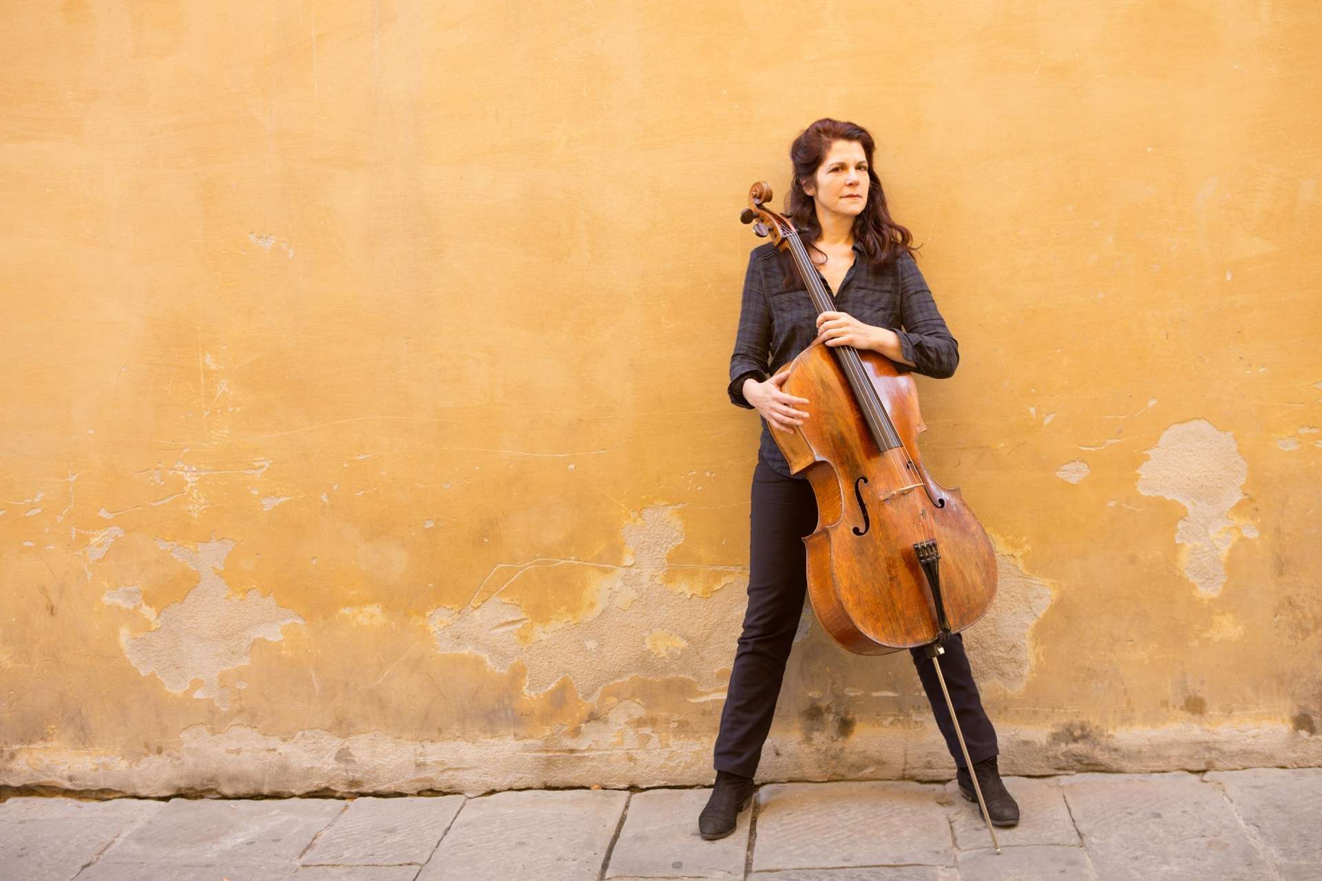 Photo de Sonia Wieder Atherton devant un mur jaune, un petit peu abîmé. Elle se tient debout avec, devant elle, son violoncelle.