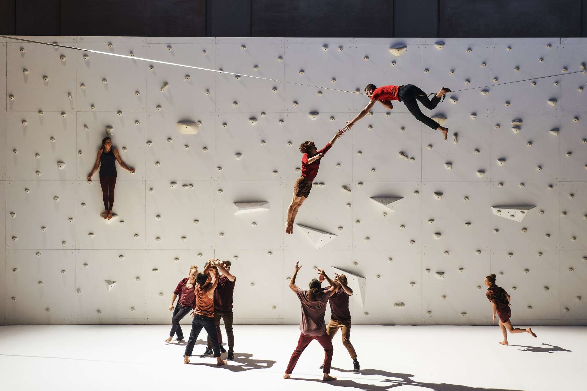 Plan large sur le spectacle avec deux groupes qui lancent une acrobate dans les airs pour essayer qu'elle rejoignent l'homme allongé plus haut sur un fil. En fond de scène, un mur d'escalade blanc. 