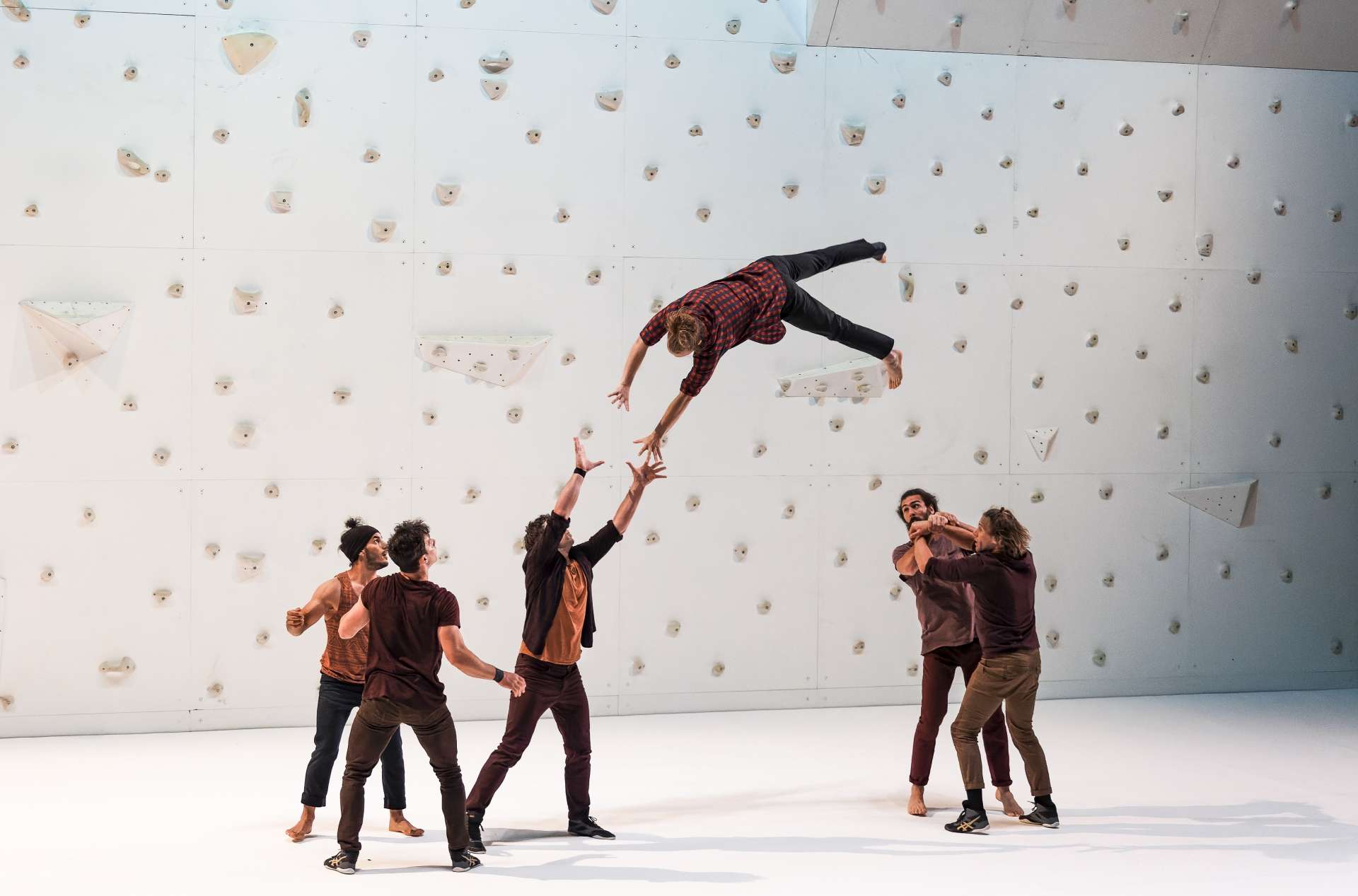 Six interprètes effectuent un porté acrobatique devant le mur d'escalade blanc situé en fond de scène.