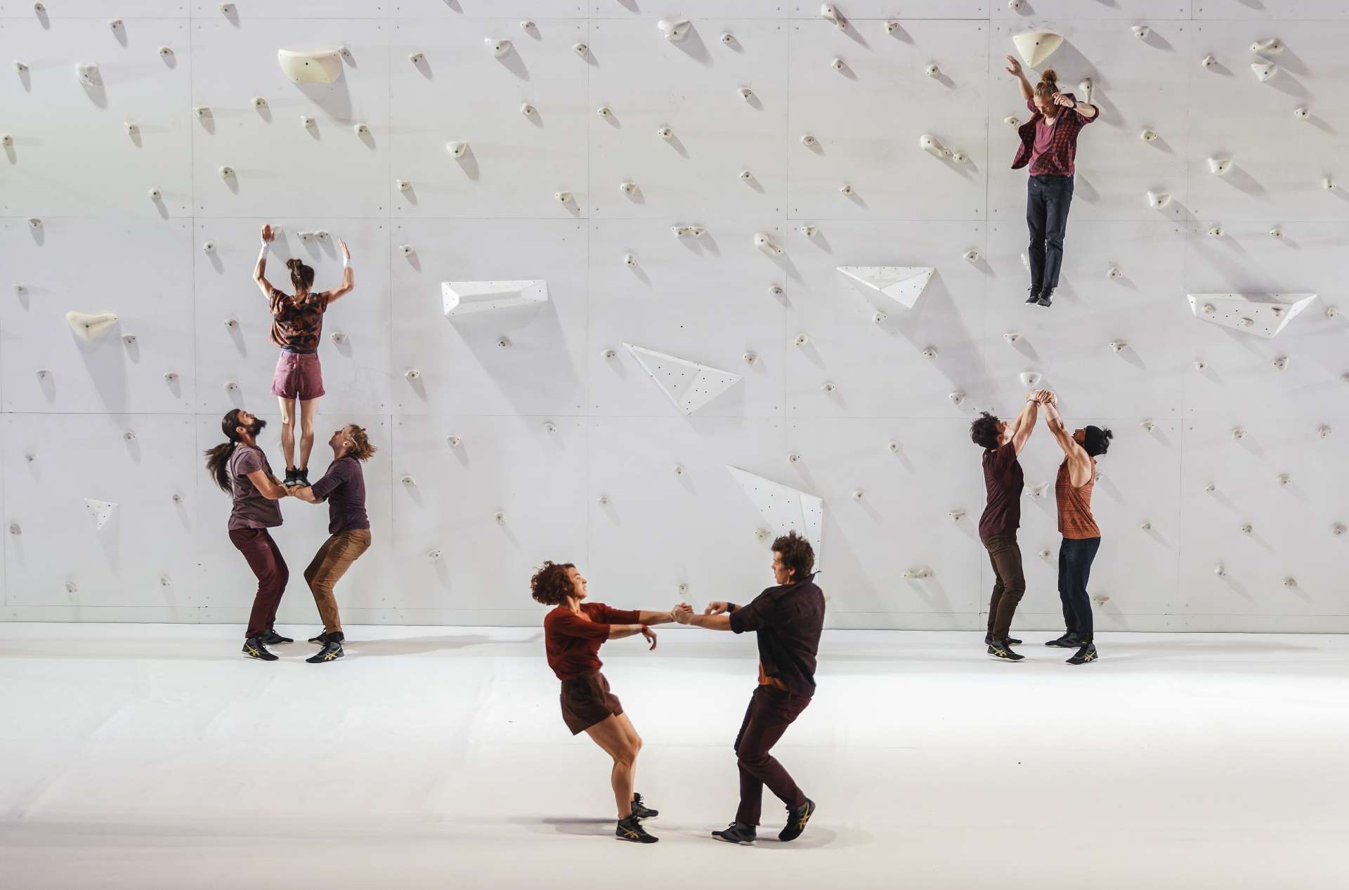 Trois groupes d'acrobates, tous en train de réaliser de portés devant le mur d'escalade blanc installé en fond de scène. 