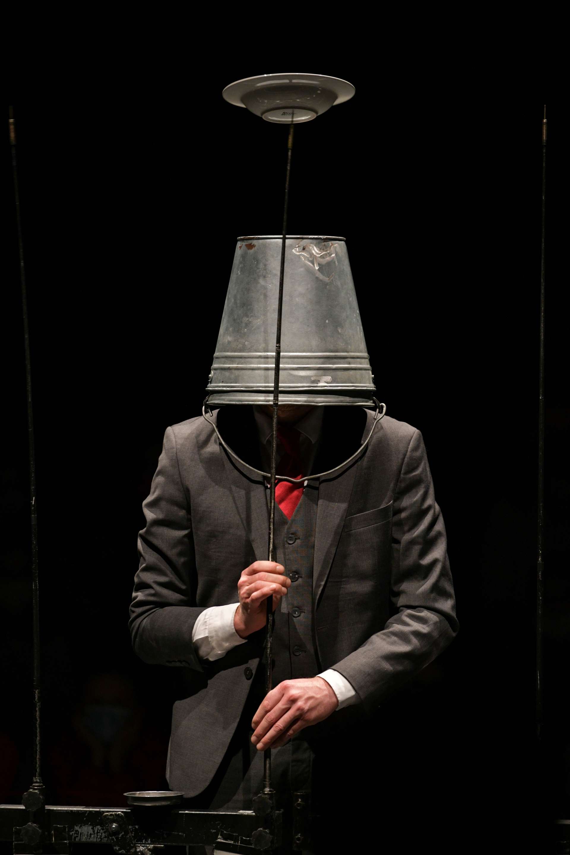 Un homme en costume gris, seau gris en métal sur la tête, tient une barre métallique avec une assiette posée en équilibre dessus. 