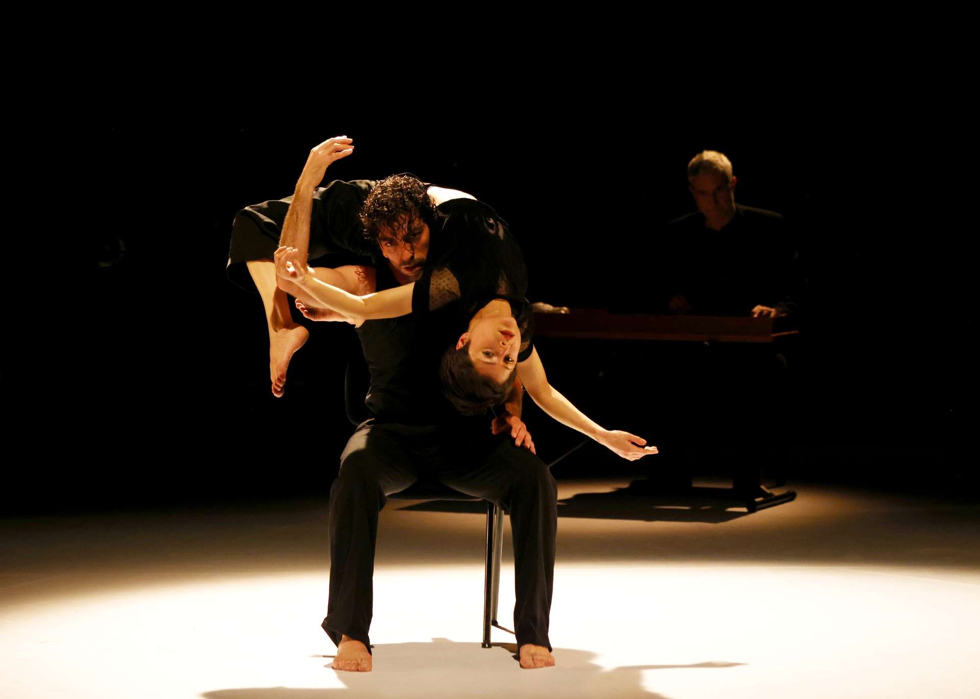 Assis sur une chaise, le danseur David Coria porte la danseuse Jann Gallois qui se contorsionne autour de son buste. 