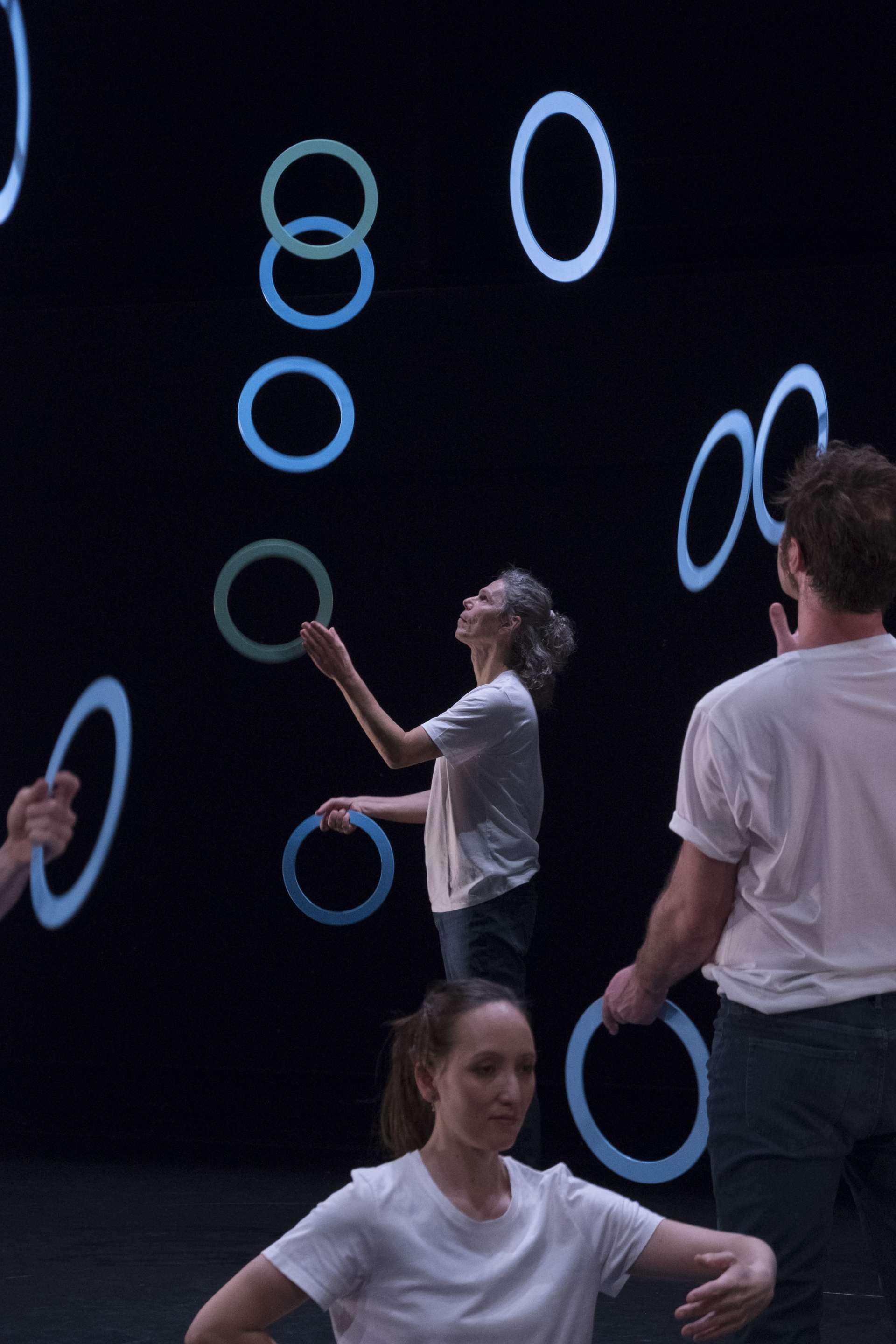 Une danseuse et un danseur interprète des mouvements de danse et de jonglage avec les cerceaux bleus au premier plan. En arrière plan, Sean Gandini fait de même. 