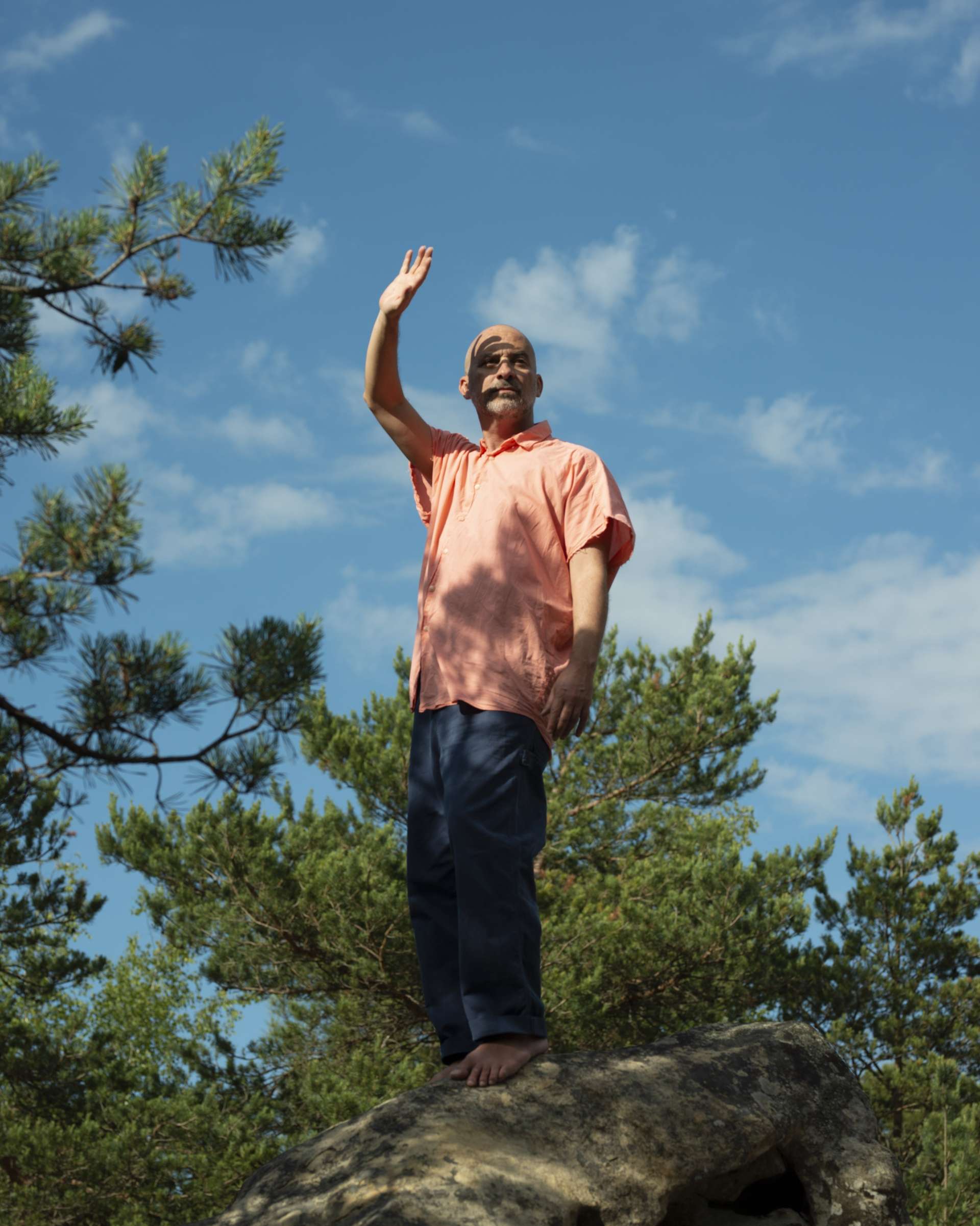 Lucas Santtana est en pleine nature, debout sur un rocher, et lève sa main droite vers le ciel bleu comme pour saluer. 