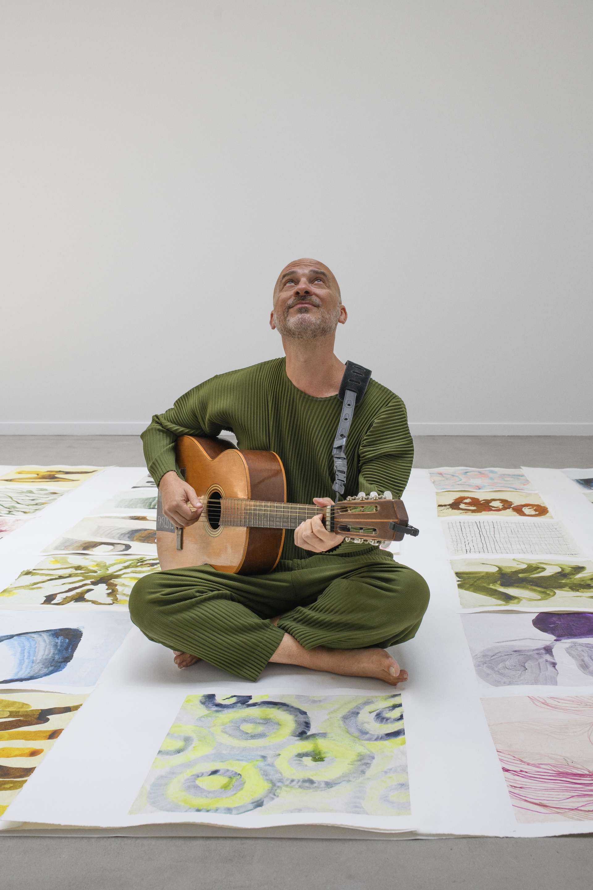 Assis en tailleur au sol sur une immense toile colorée, Lucas Santtana, guitare à la main, regarde le ciel. 