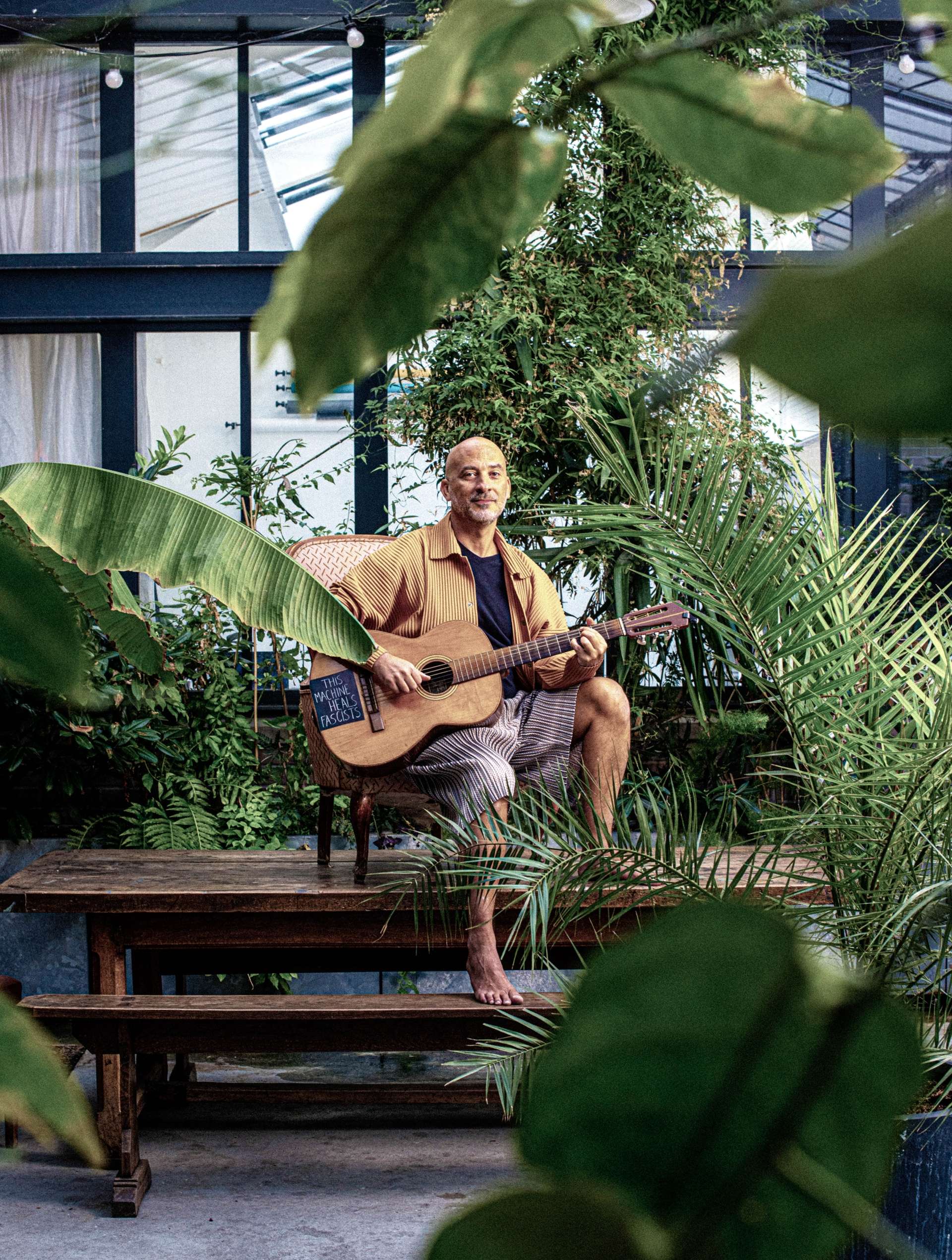 Assis sur une table en bois, guitare à la main, Lucas Santtana pose face à l'objectif au milieu d'une végétation luxuriante de type jungle urbaine d'intérieur. 