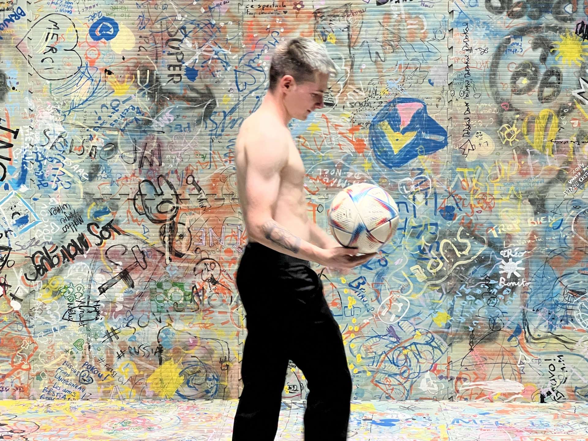 Paul Molina, torse nu et ballon de foot à la main, positionné de profil devant un mur de street art. 