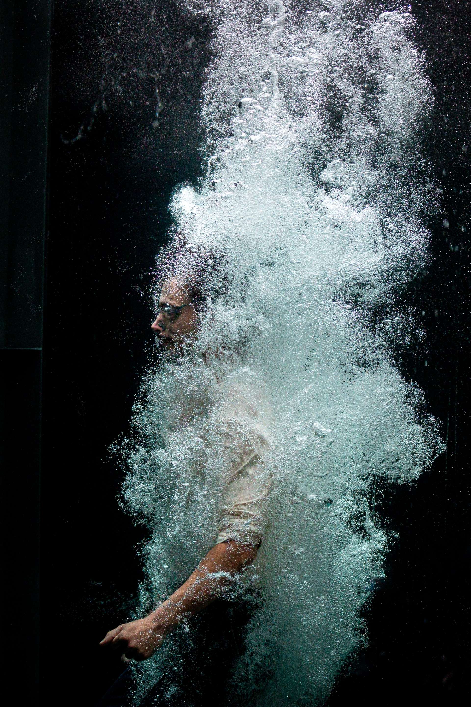 Un apnéiste habillé, de profil, sous l'eau et entouré d'un rideau de fines bulles. 