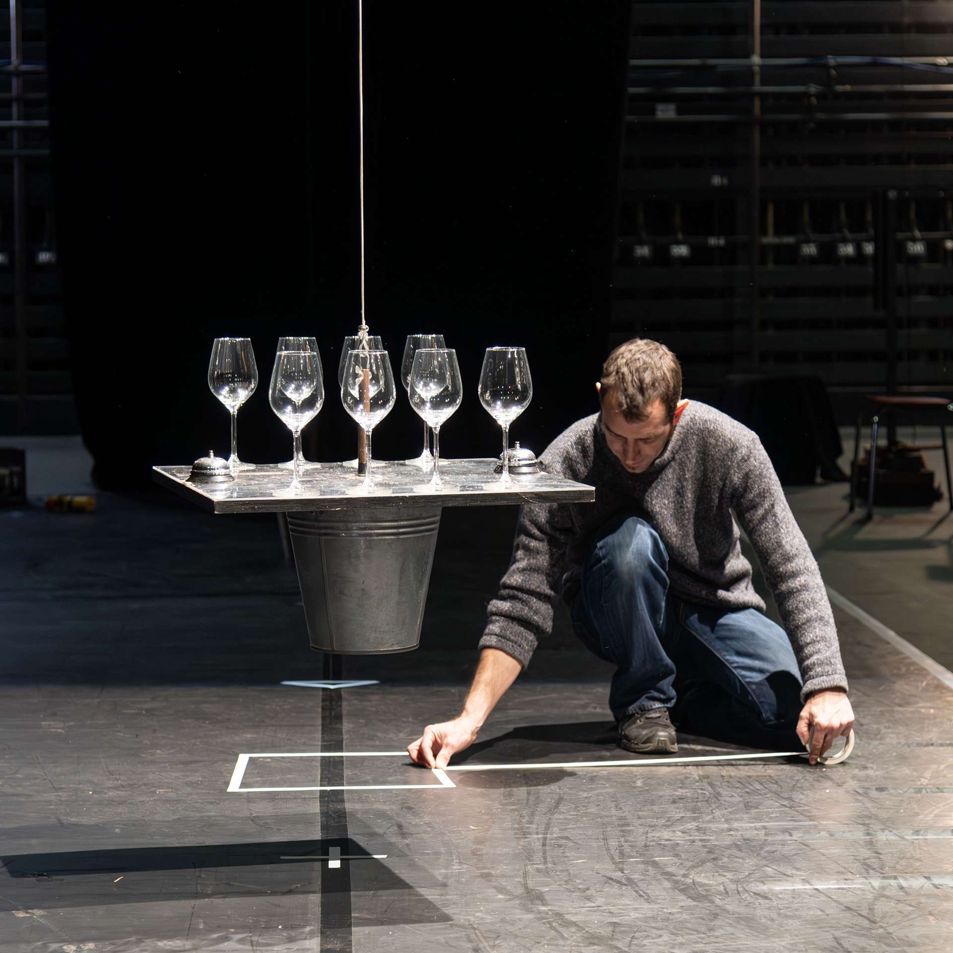 Un technicien pose du gaffer blanc sur le sol pour faire un repère. A côté de lui, des verres sont posés sur une petite plateforme suspendue. 