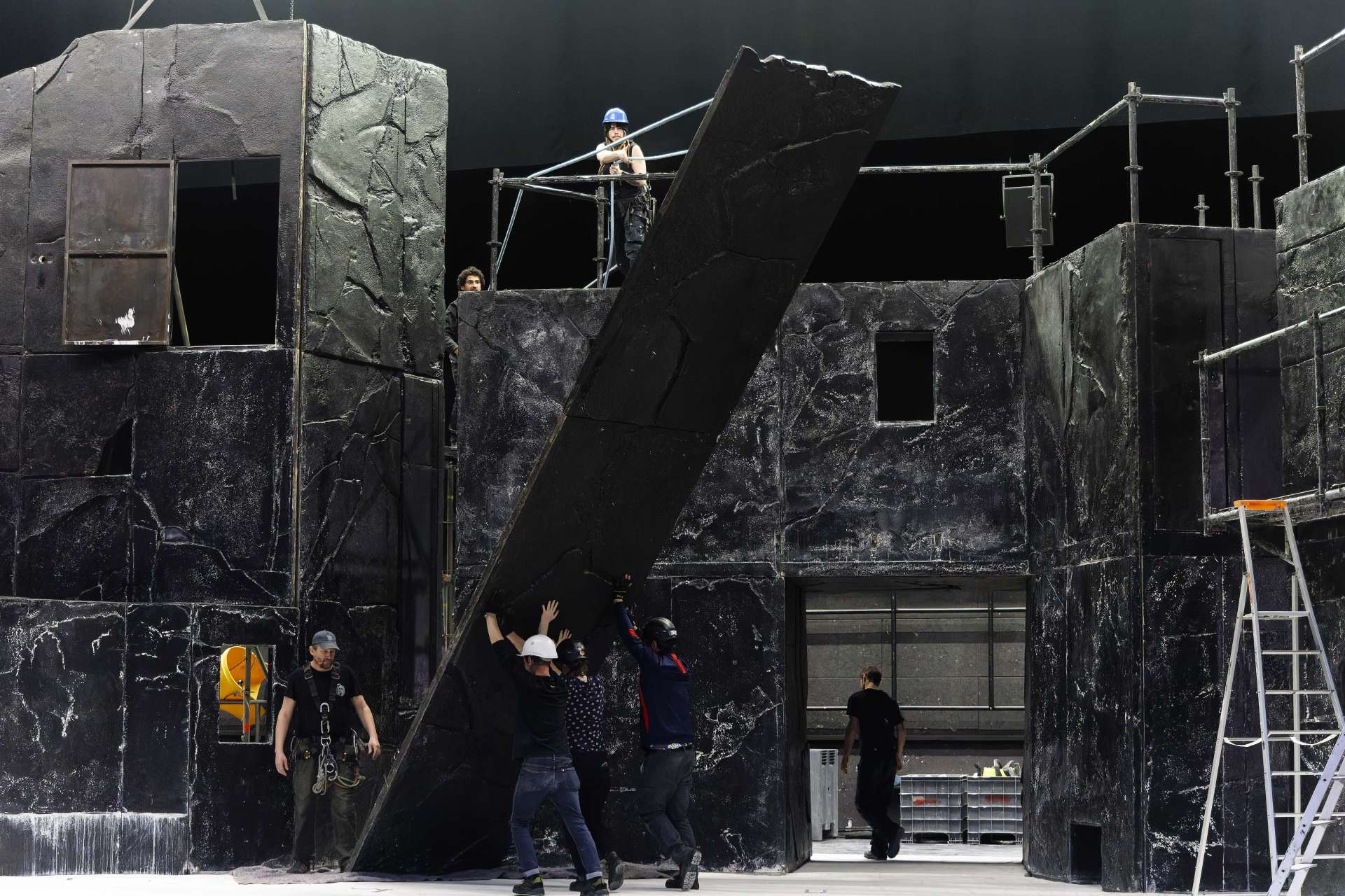 Plusieurs techniciens portant des casques montent un décor noir de plusieurs mètres de haut 