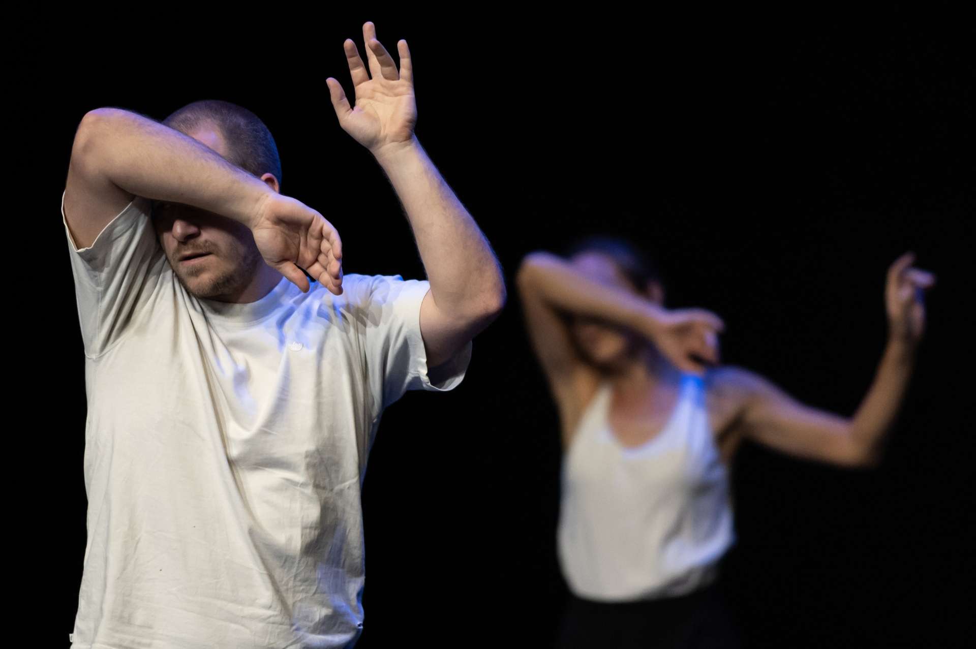 Caché derrière son coude, un interprète danse au premier plan. Au second, une interprète a la même posture. 