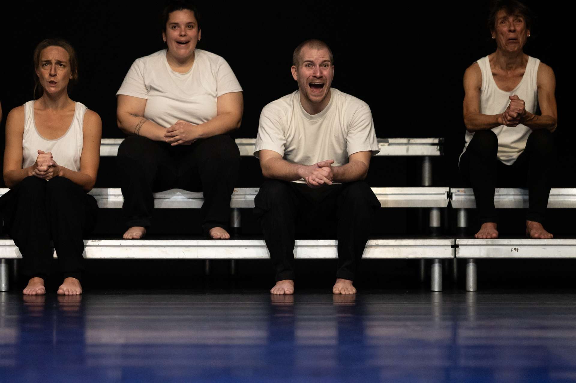 Trois femmes et un homme sont assis dans un gradin sommaire. Pantalon noir, débardeur blanc et chacun une expression singulière sur le visage. 