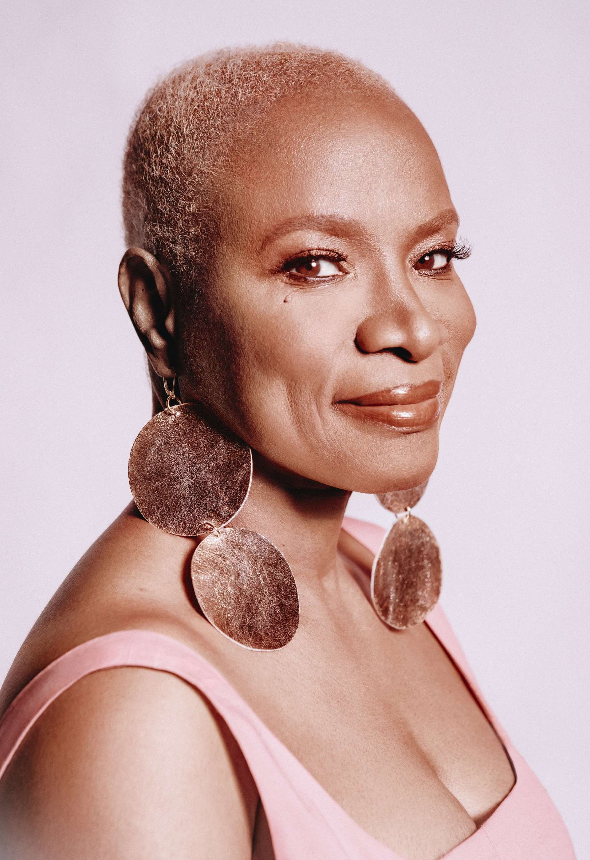 Photo portrait de trois quart de l'artiste Angélique Kidjo. Les couelurs sont douces, elle porte un débardeur rosse et des immenses boucles d'oreille rosées formées de deux ronds de cuir. 