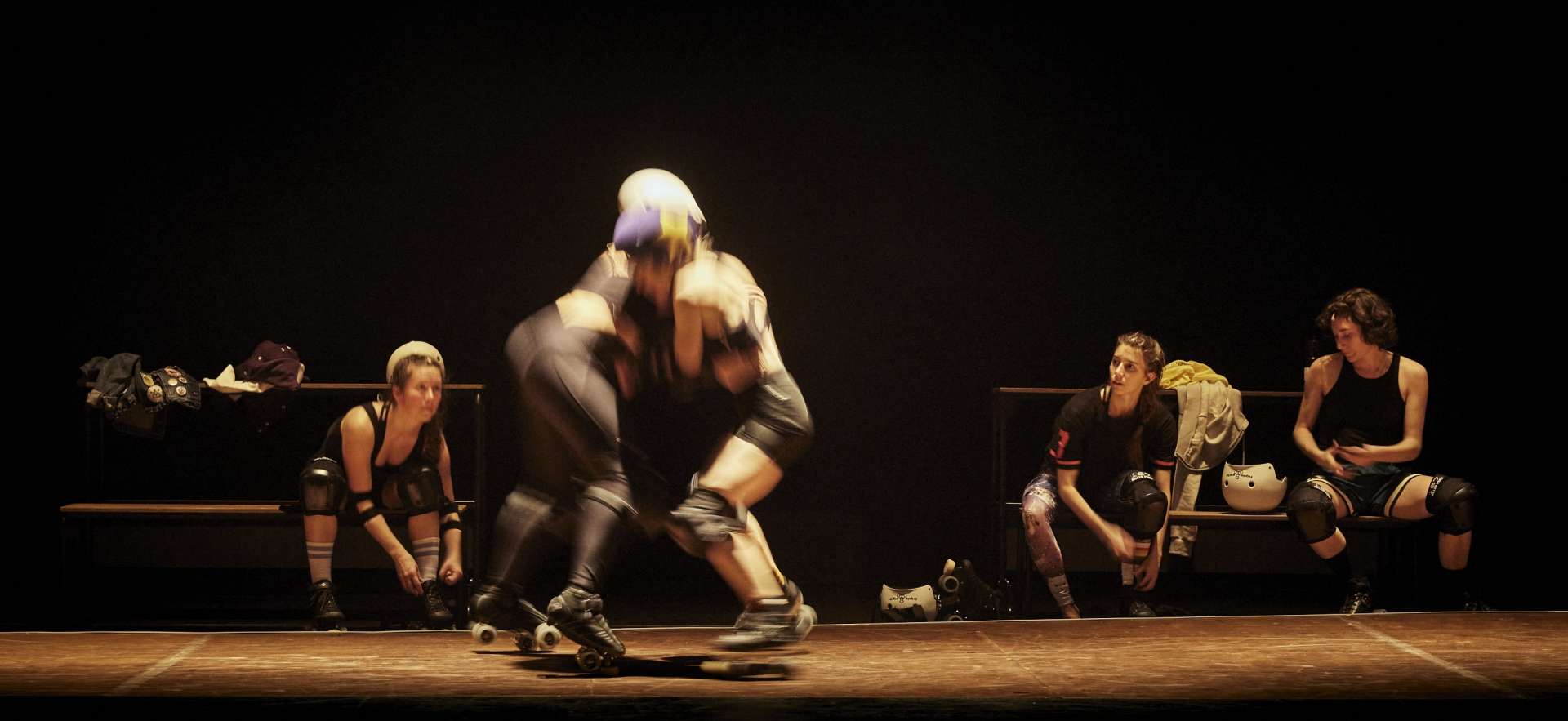 Deux interprètes en roller sont en face à face sur une piste en bois. Au second plan, trois interprètes enfilent leurs rollers.