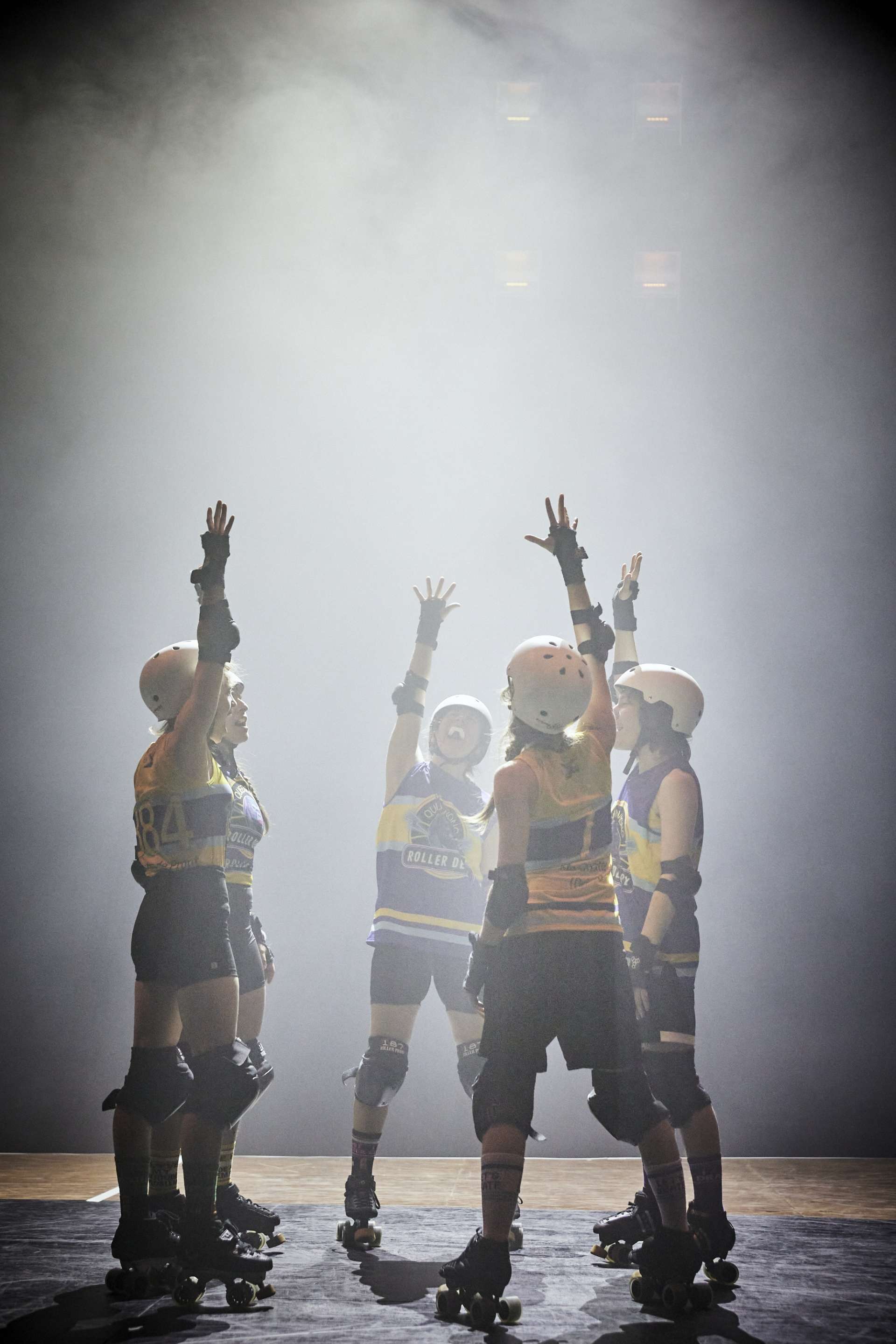 Cinq interprètes sont en cercle. Elles lèvent une main en l'air et porte un équipe de roller derby : casque, maillot jaune et bleu, protections et patins à roulettes. 