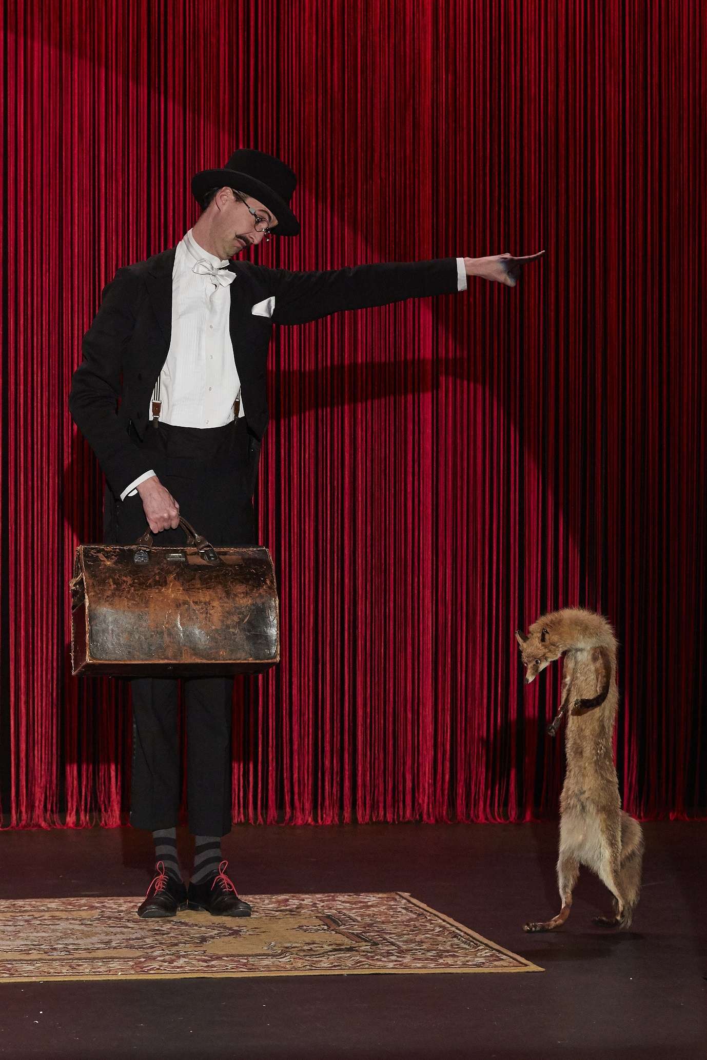 Le magicien Kosma est vêtu d'un beau costume et d'un chapeau haut-de-forme. Il a une valise à la main et semble pointer la sortie à son assistant renard prénommé Goupil. 