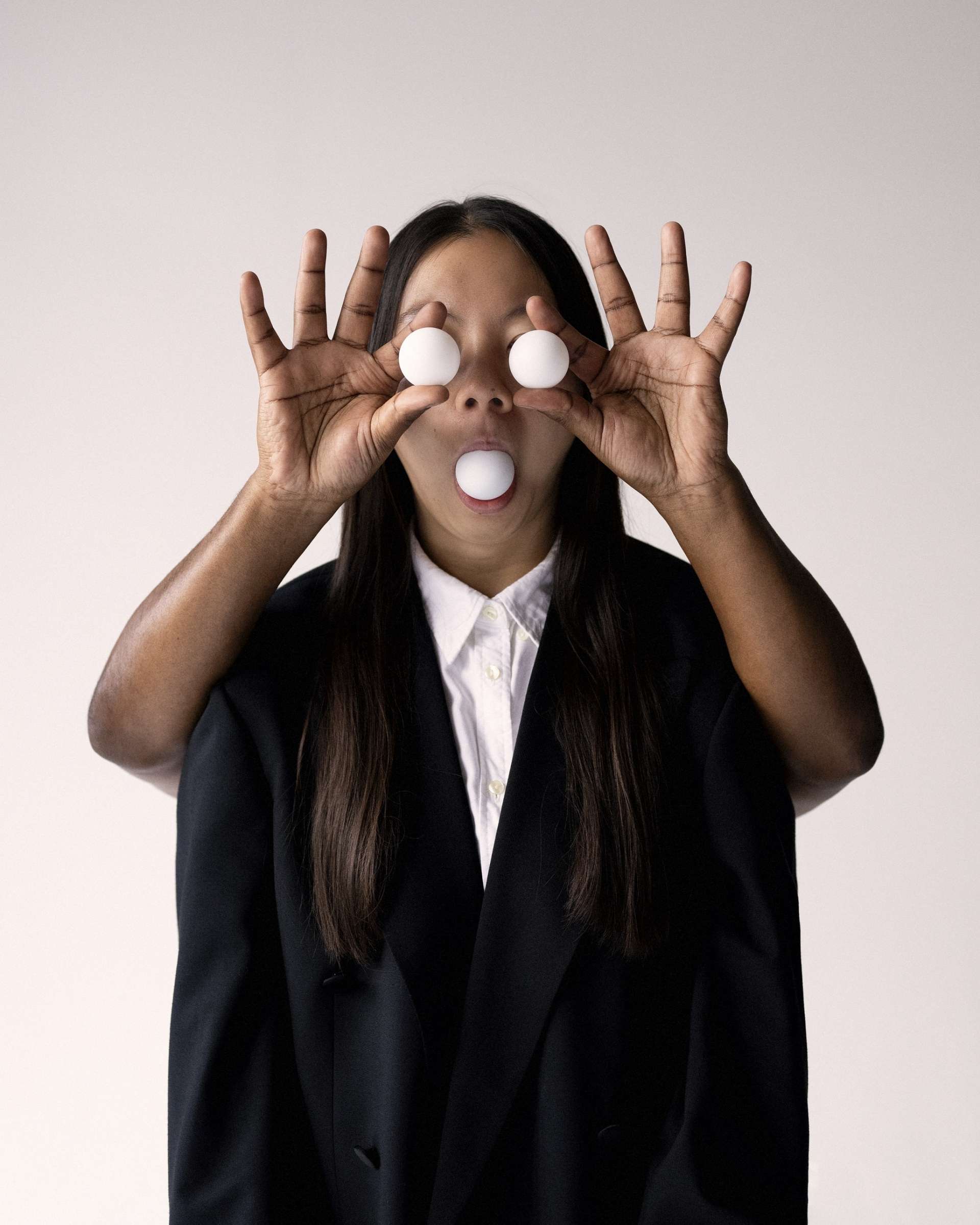 Portrait d'une femme en costume noir et chemise blanche. Ses yeux et sa bouche sont remplacés par des balles de ping-pong. Elles sont tenues par des mains qui ne sont pas les siennes mais semblent l'être. 