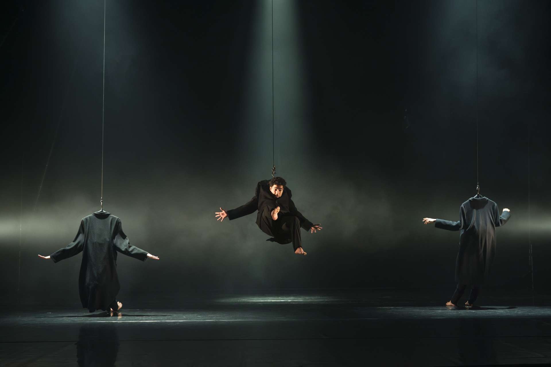 Trois acrobates en costard sont accrochés dans les airs et dansent dans la pénombre. 