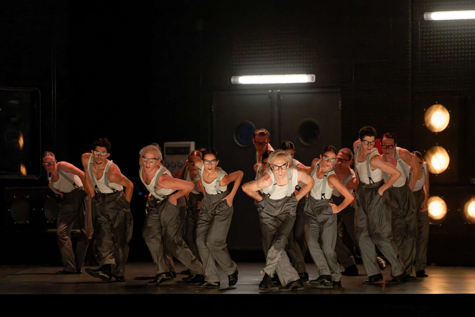 Photo de groupe des interprètes du ballet qui dansent avec pour uniforme un pantalon gris, des bretelles, un débardeur blanc et des lunettes. 