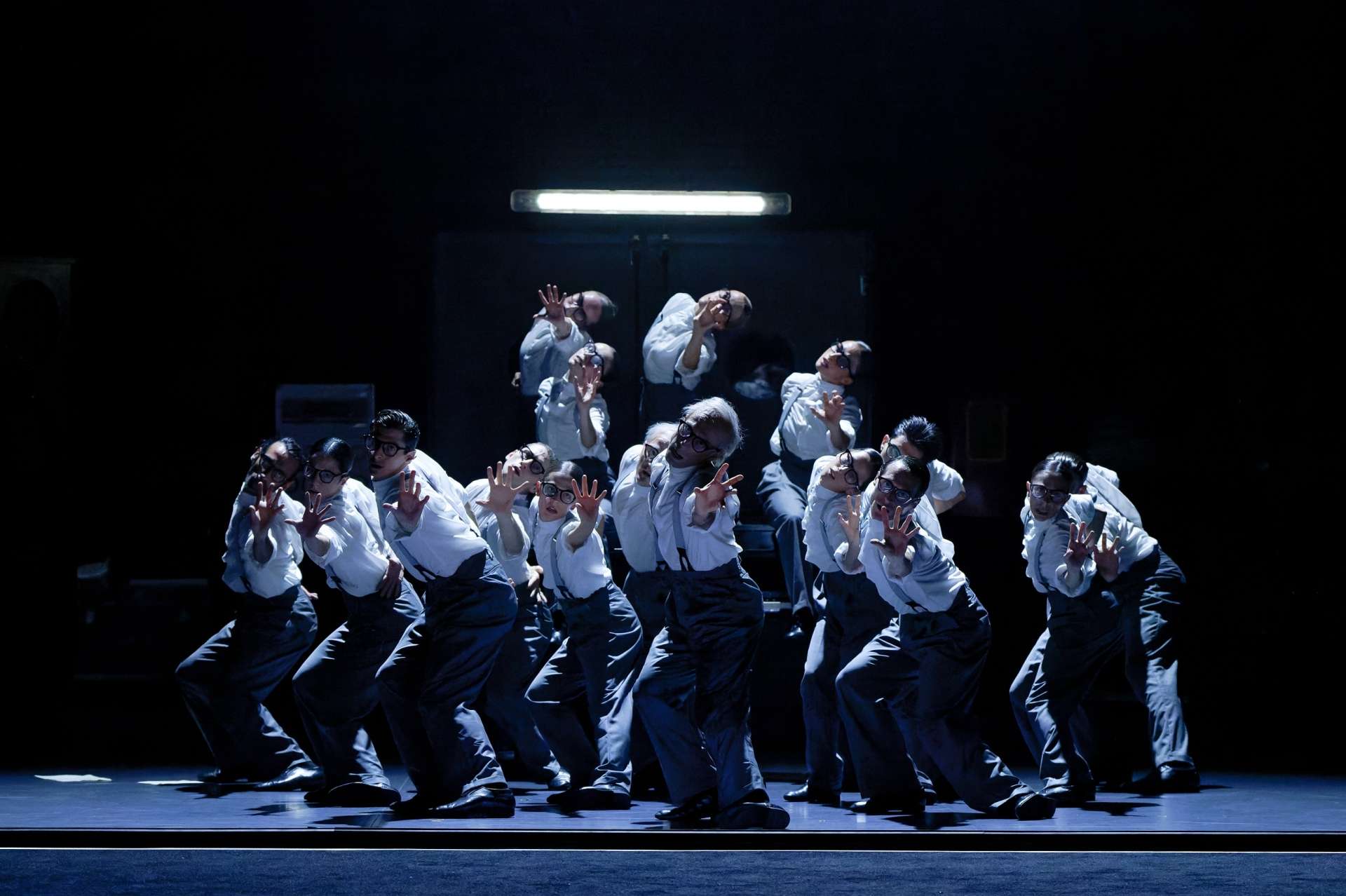 Photo de groupe des interprètes du ballet qui dansent dans une lumière bleutée avec pour uniforme un pantalon gris, des bretelles, un débardeur blanc et des lunettes. 